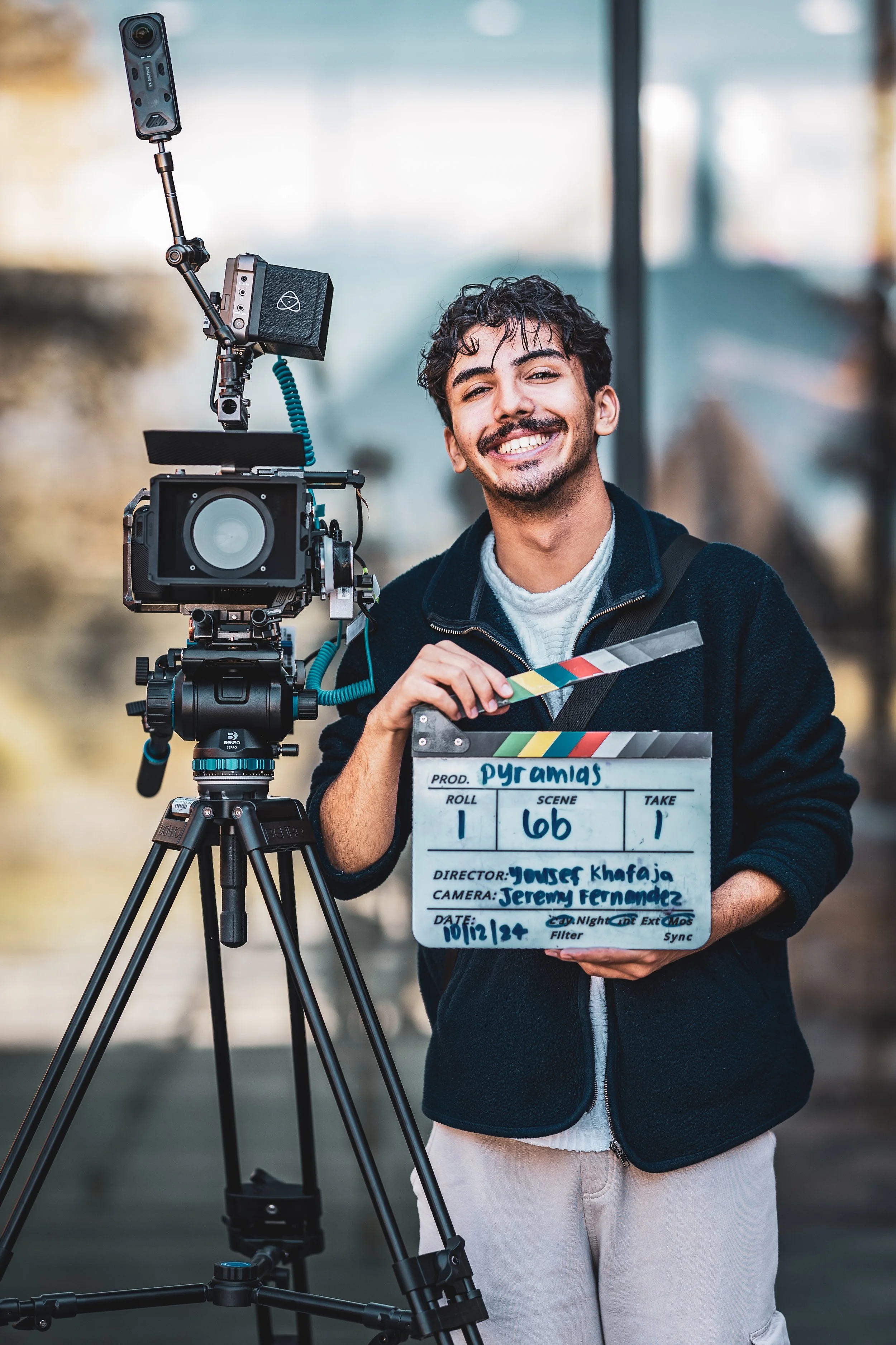 A young man holding a film clapboard standing next to professional filming equipment in an outdoor setting.