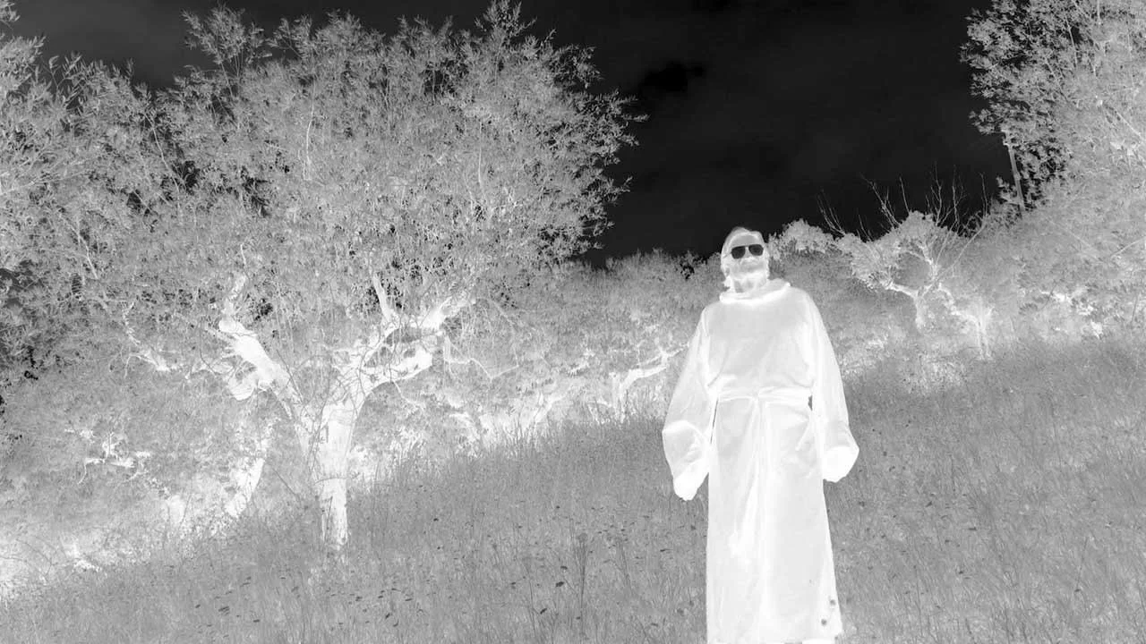 A person wearing sunglasses and a long white robe, standing on grassy terrain with trees in the background, appearing in a negative image.
