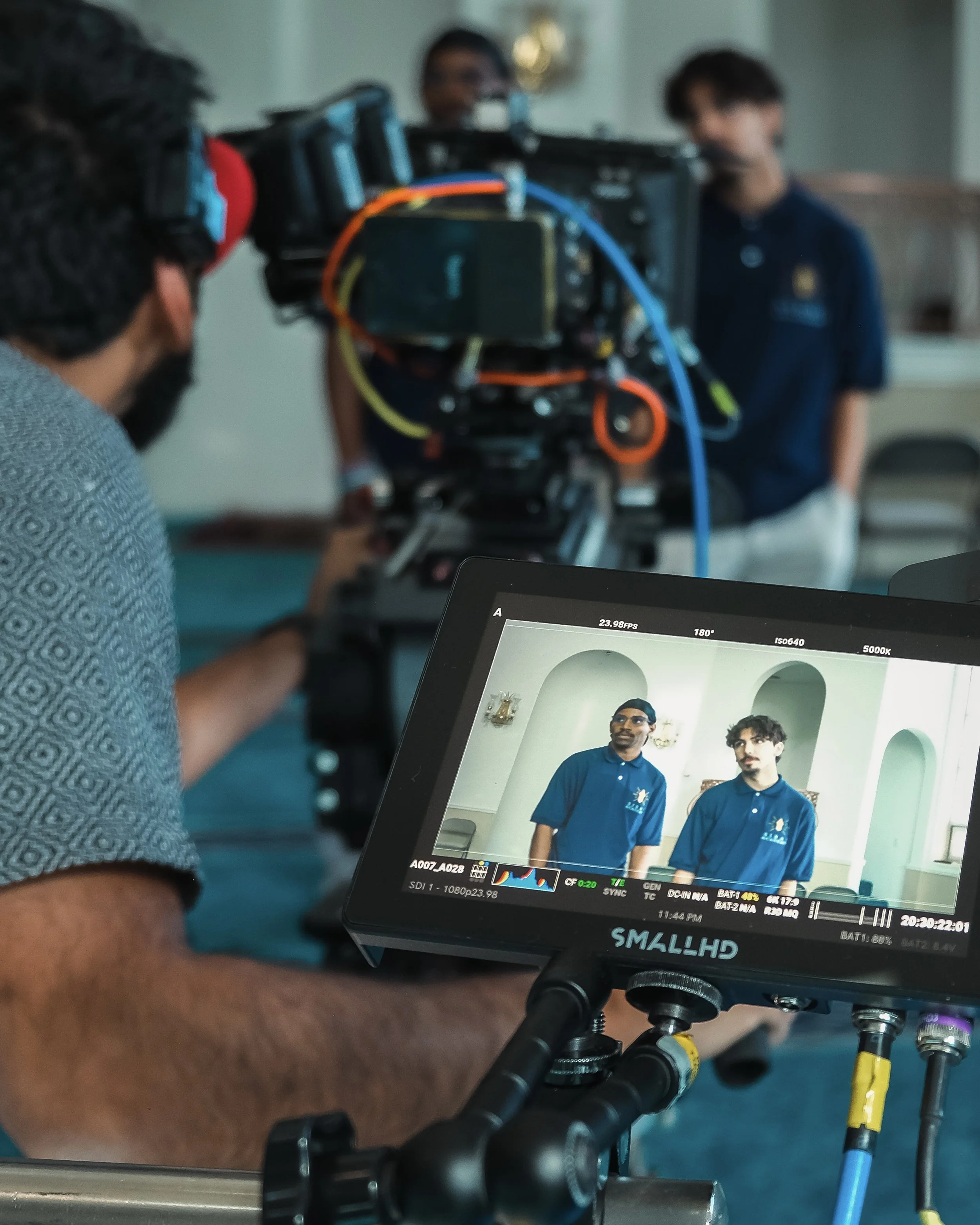 Two men are on camera in front of a turquoise wall with arched windows, wearing matching blue polo shirts with a logo, looking slightly off-camera.