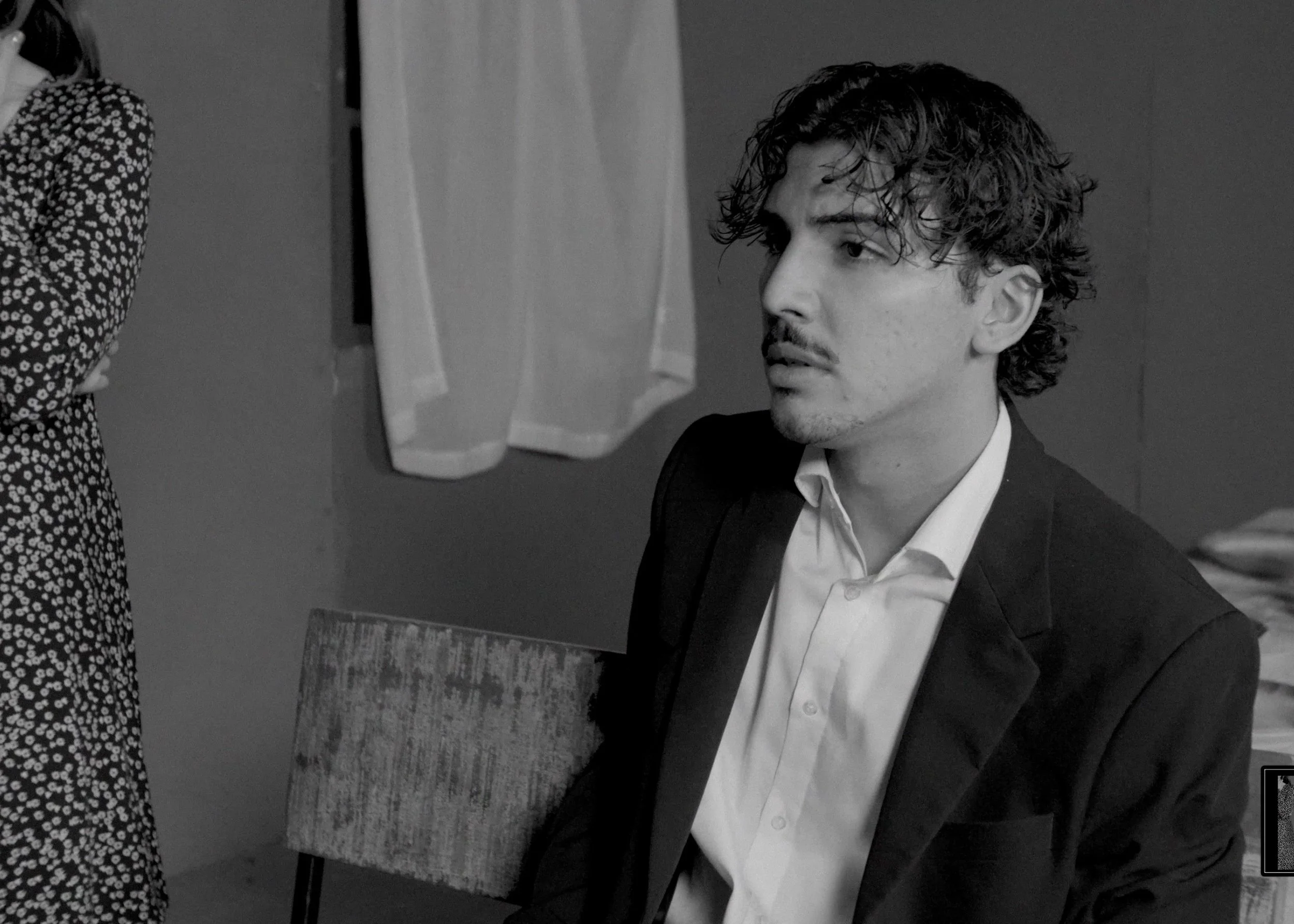 Black and white photo of a young man with curly hair, wearing a suit and white shirt, sitting and looking to his right, with a woman partially visible on the left, in an indoor setting with a chair and towels hanging in the background.