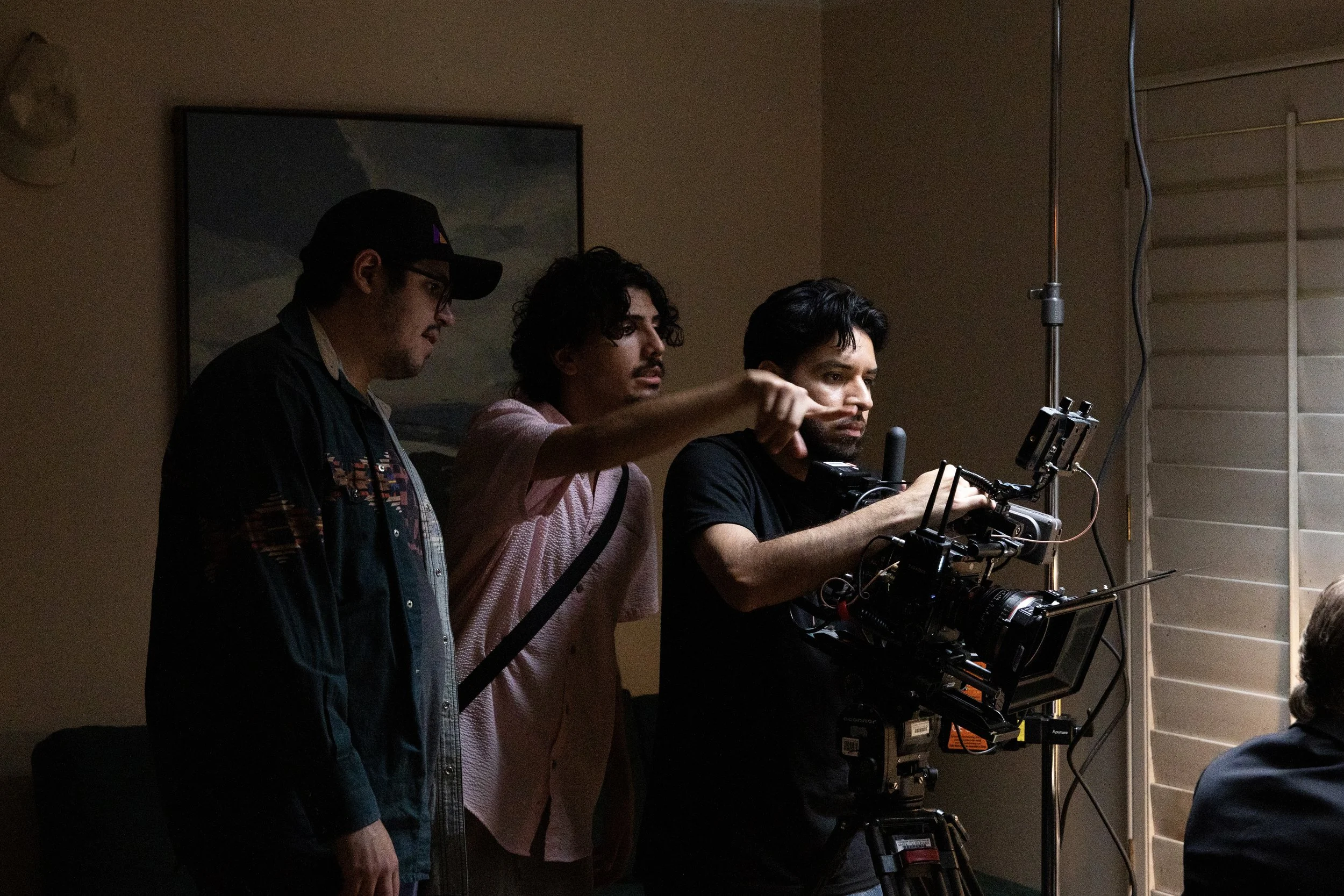 Three men working on a film set with a camera, with blinds on a window and a landscape painting on the wall in the background.