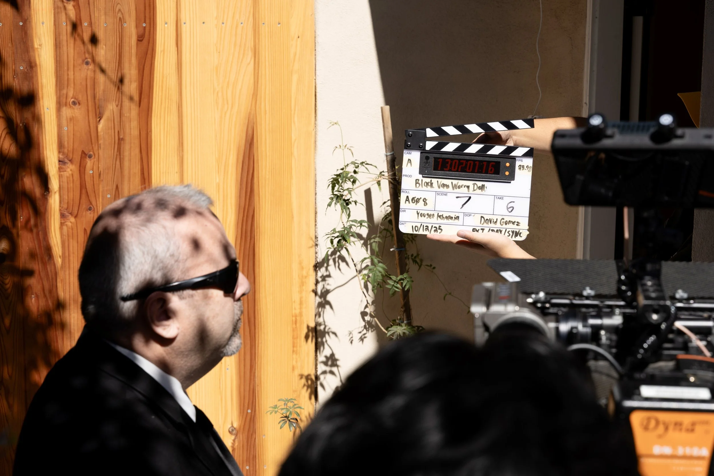 A man wearing sunglasses is sitting in front of a wooden wall while a clapperboard is being held above him. The clapperboard has details written on it and is part of a film or video production scene.