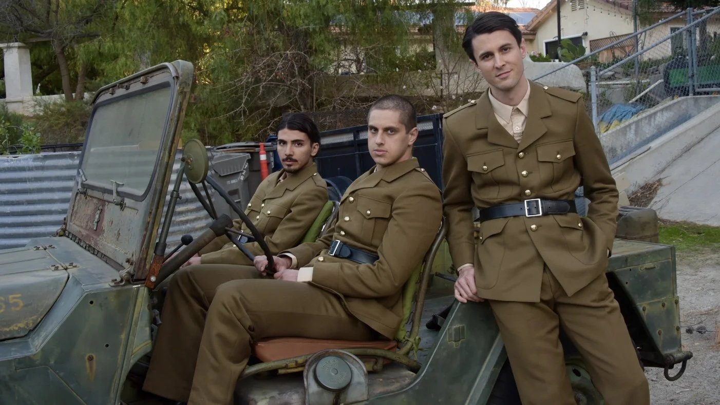 Three men in military uniforms sitting and standing on an old military vehicle outdoors with trees and houses in the background.