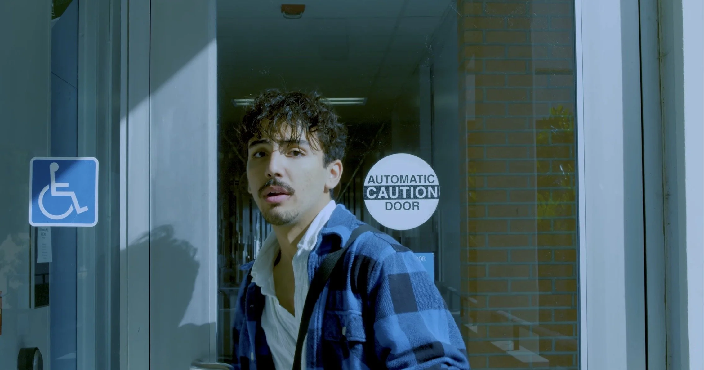 A young man with dark curly hair and a mustache standing outside a glass door with a sign that says "Automatic Caution Door," and a blue wheelchair accessibility sign on the door.