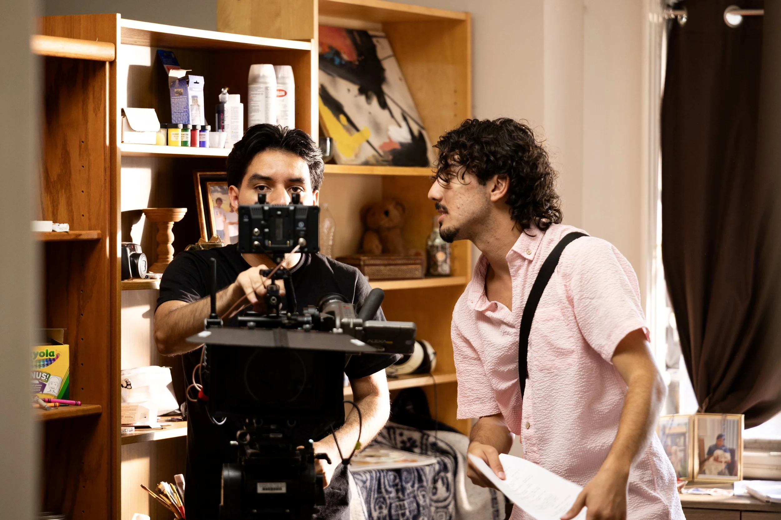 Two men on a film set, one operating a camera and the other holding a script, in a room with wooden shelves and personal items.