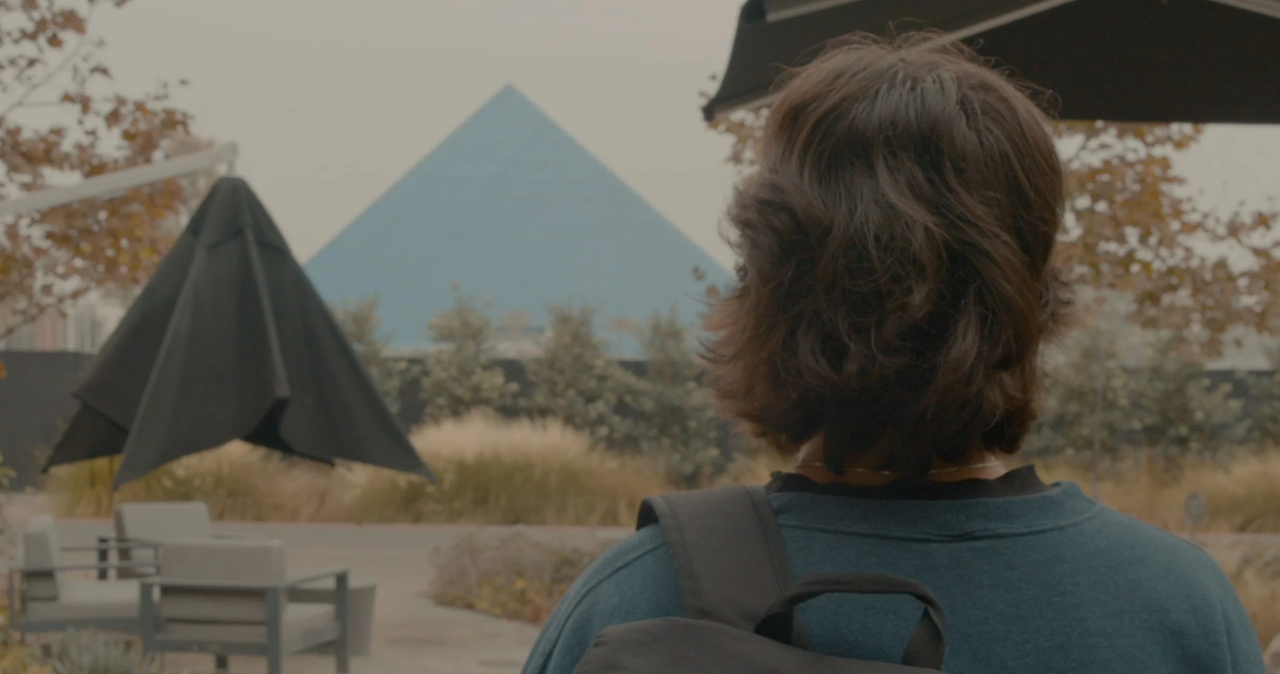 A person with curly brown hair is seen from behind, standing outdoors with a backpack, looking towards a blue pyramid-shaped building in the distance, with outdoor seating and umbrellas around.