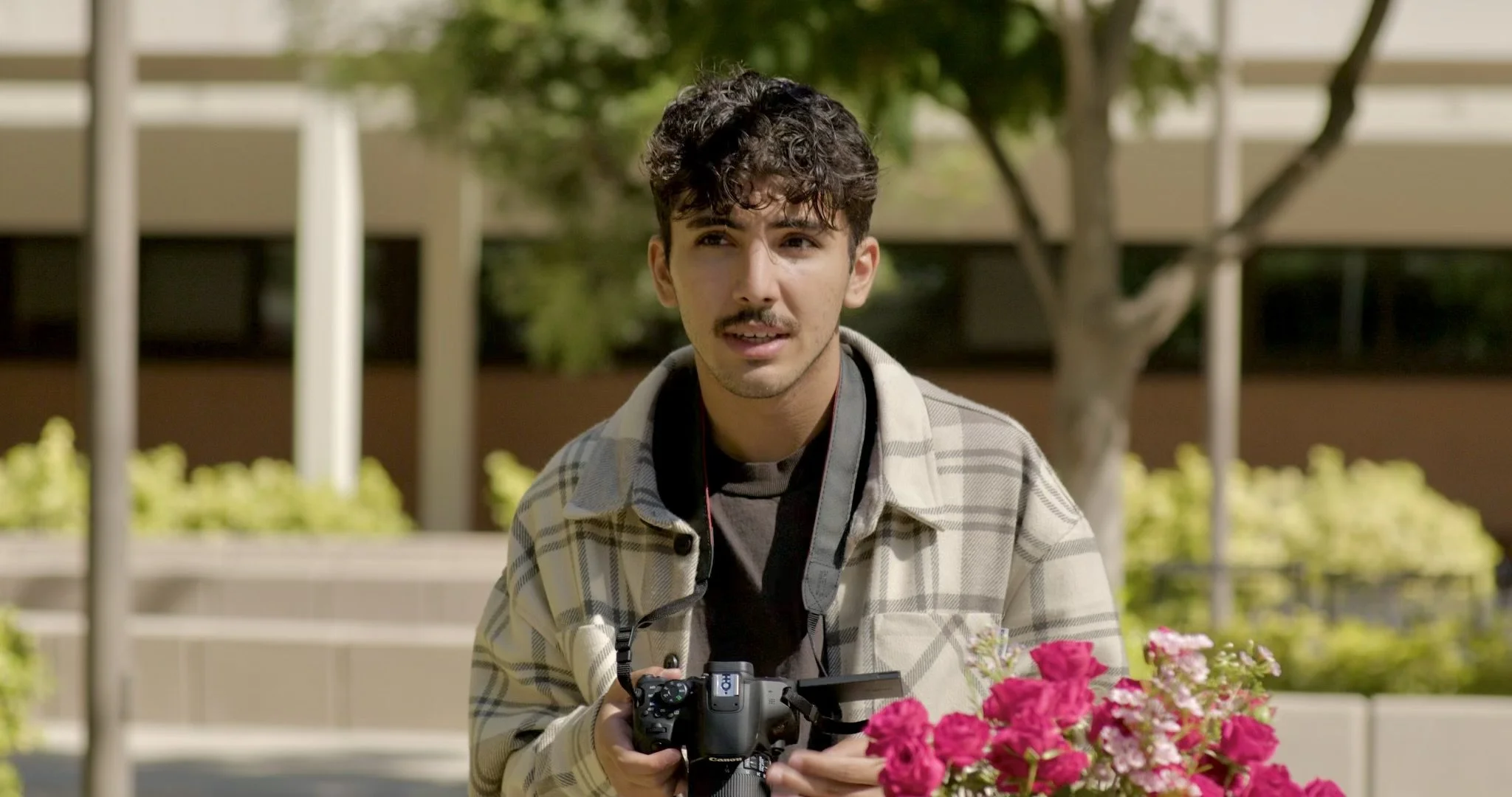 A young man with curly dark hair and a mustache holding a camera, standing outdoors near pink flowers and trees, with a school or office building in the background.
