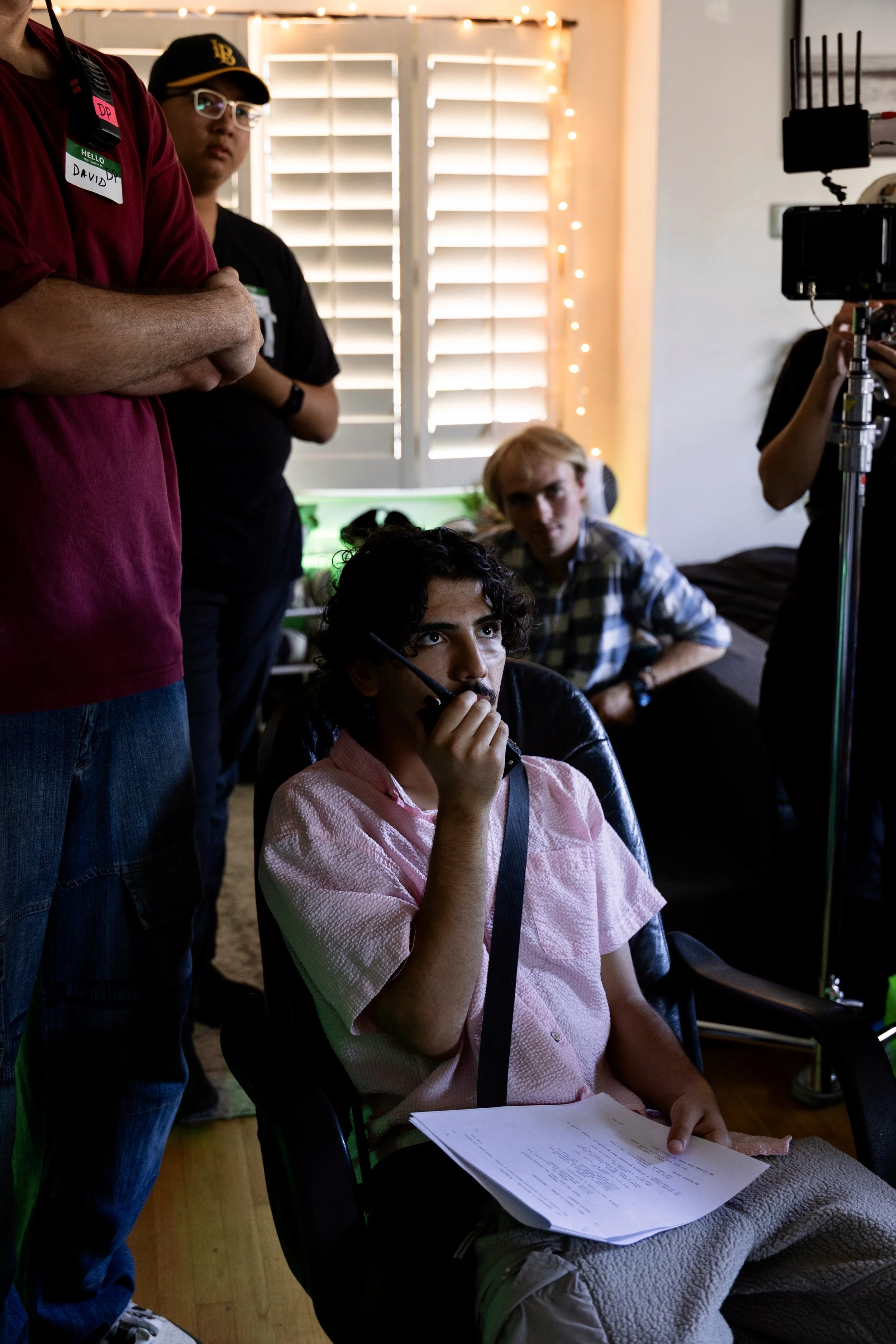 A group of five people involved in a film or video production inside a room with a window with blinds and string lights. One person is sitting in a chair holding a walkie-talkie and papers, while others stand and watch. There is filming equipment visible.