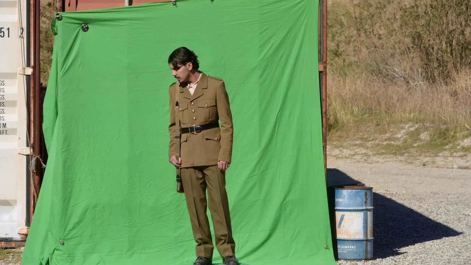 A man in a 1940s military uniform standing in front of a green screen outdoors, with a rural area and a barrel in the background.