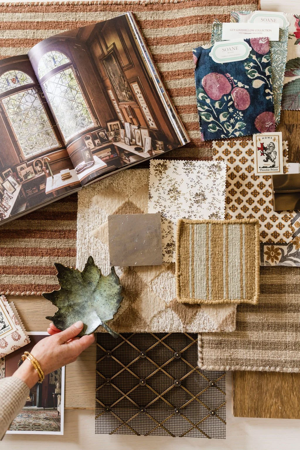 Interior room decor with fabric swatches, wallpaper samples, a decorative leaf-shaped dish, a book open to a room with large stained glass windows, and various design accessories.