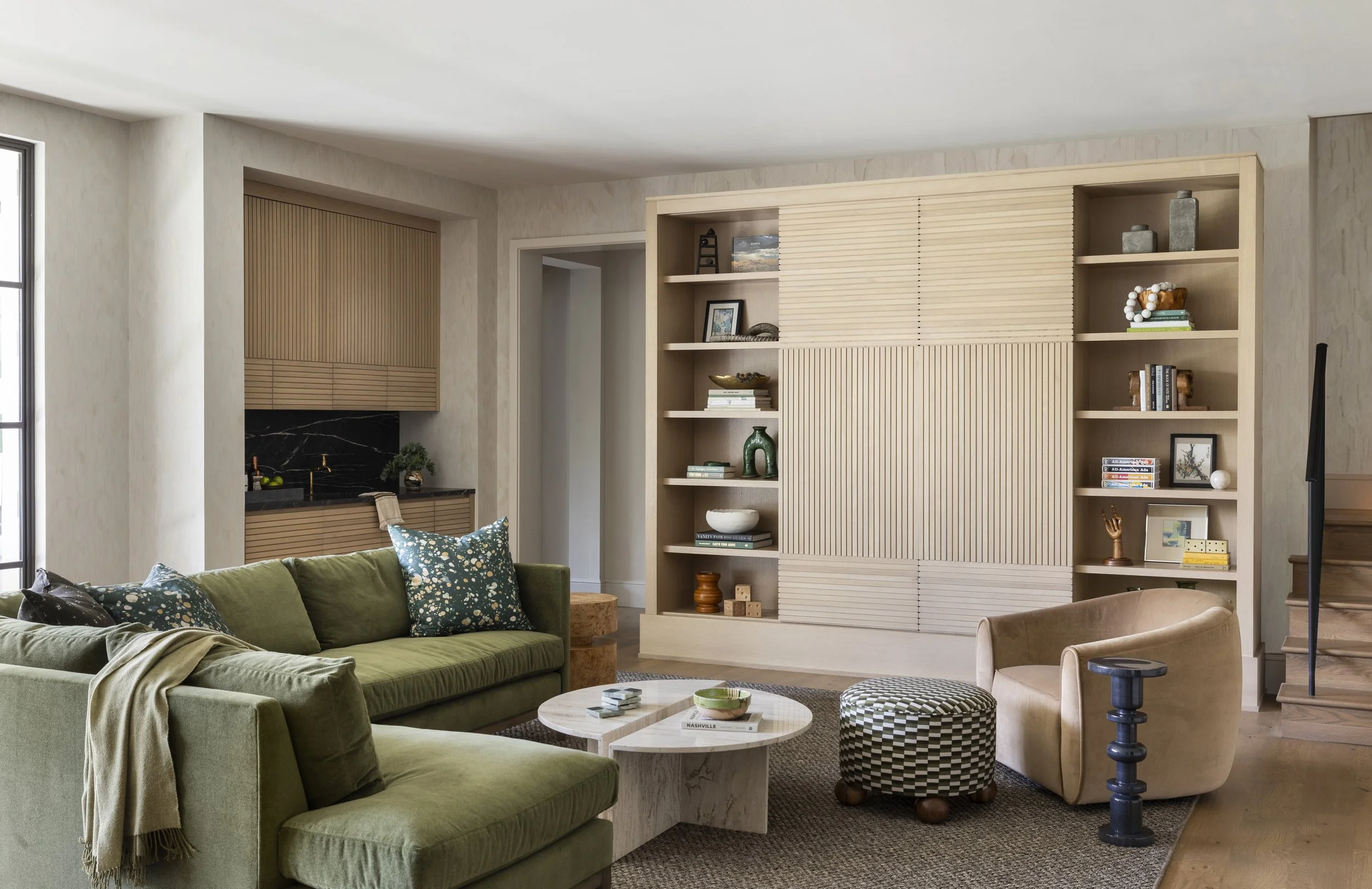 Living room with green sofa, beige armchair, wooden shelving with books and decorations, and a black and white checkered pouf.