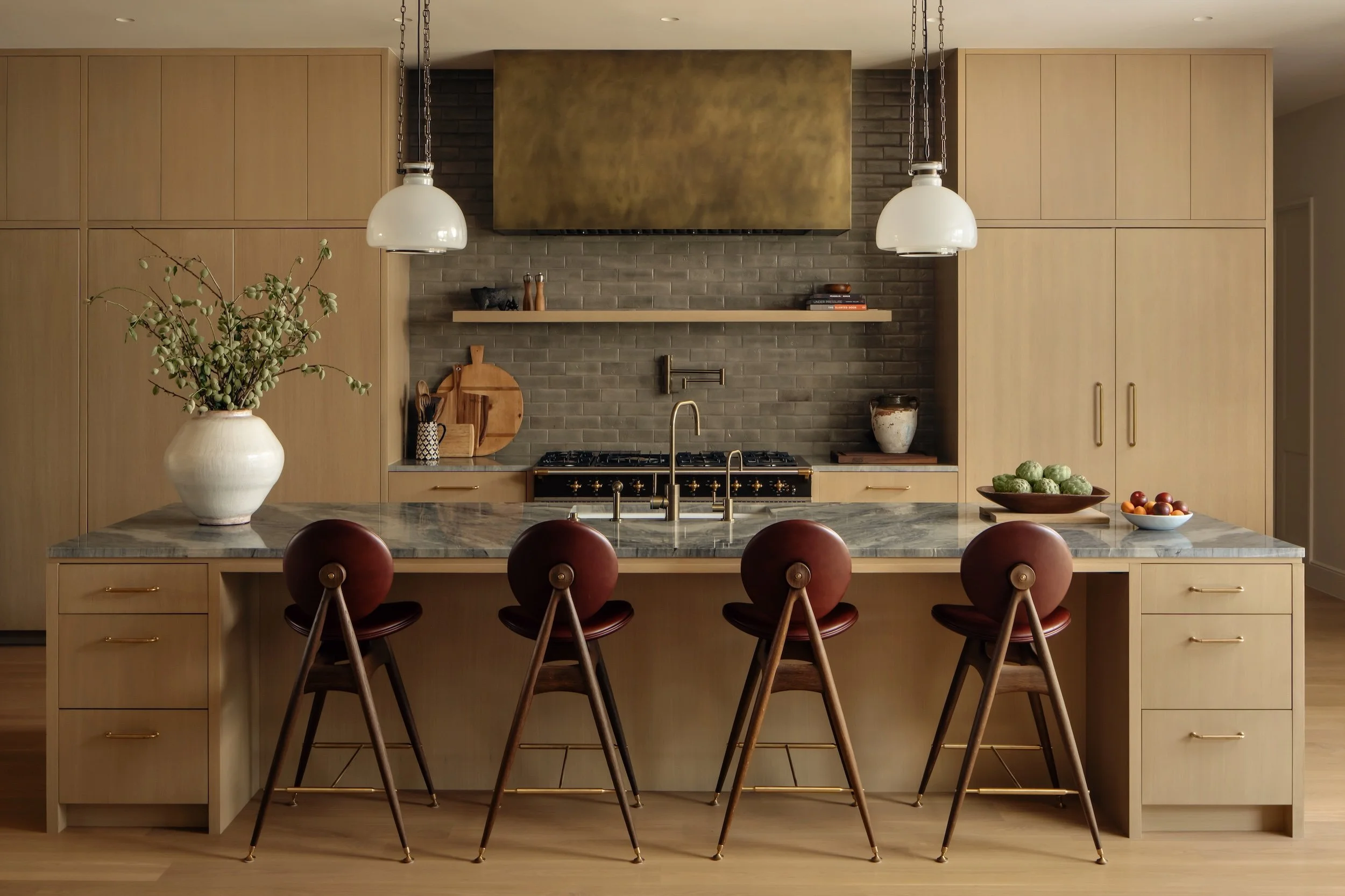 Modern kitchen with a large island, marble countertop, four red-brown bar stools, light wood cabinetry, a stove with a gold range hood, gray tiled backsplash, and decorative items including a large white vase with branches, bowls of fruit, and shelve
