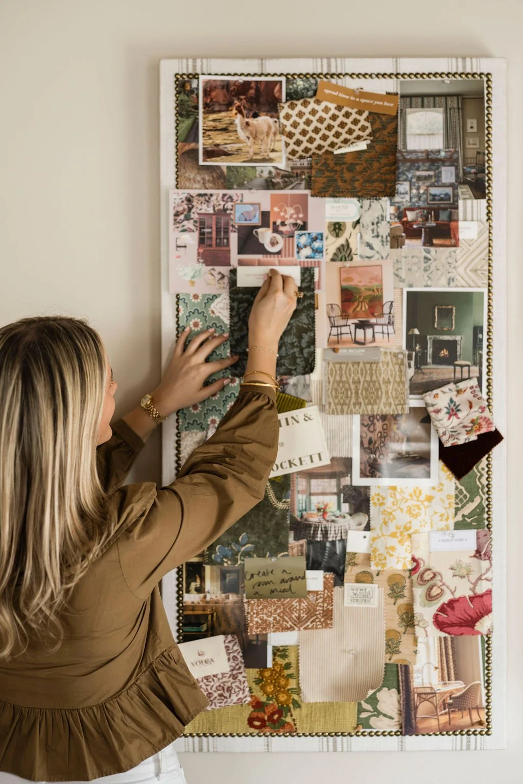 A woman working on a mood board or inspiration wall with fabric swatches, photos, and notes in a room with beige walls.