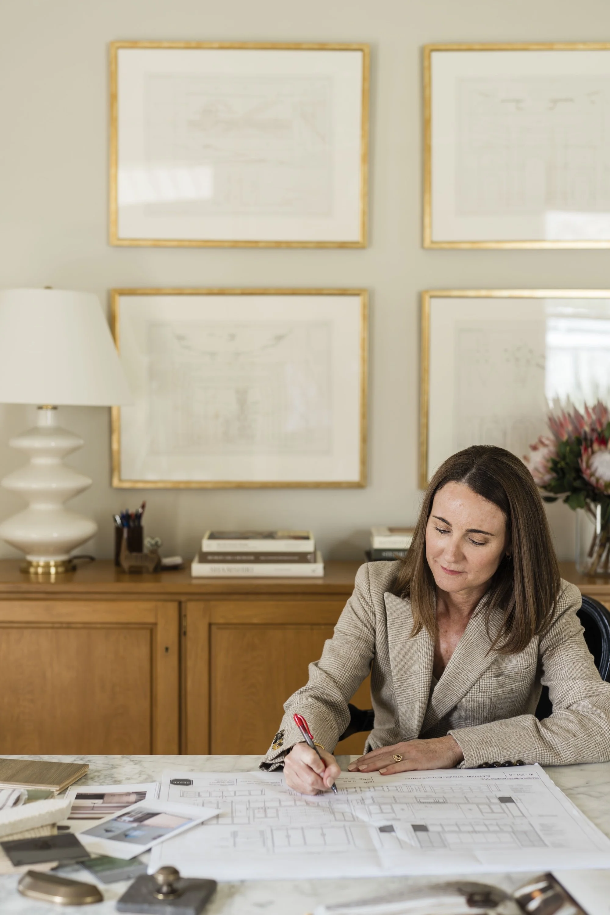 Woman in beige blazer working on architectural plans at a marble table in an office with framed blueprints on the wall, a white lamp, and a bouquet of pink flowers in a vase.