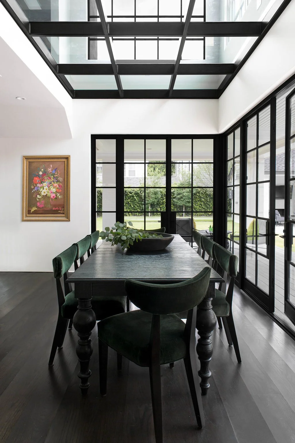 A bright white kitchen with dark hardwood floors, white cabinets, and a marble countertop. There are some vegetables on the counter, a vase with pink flowers, and decorative white vases on open shelves.