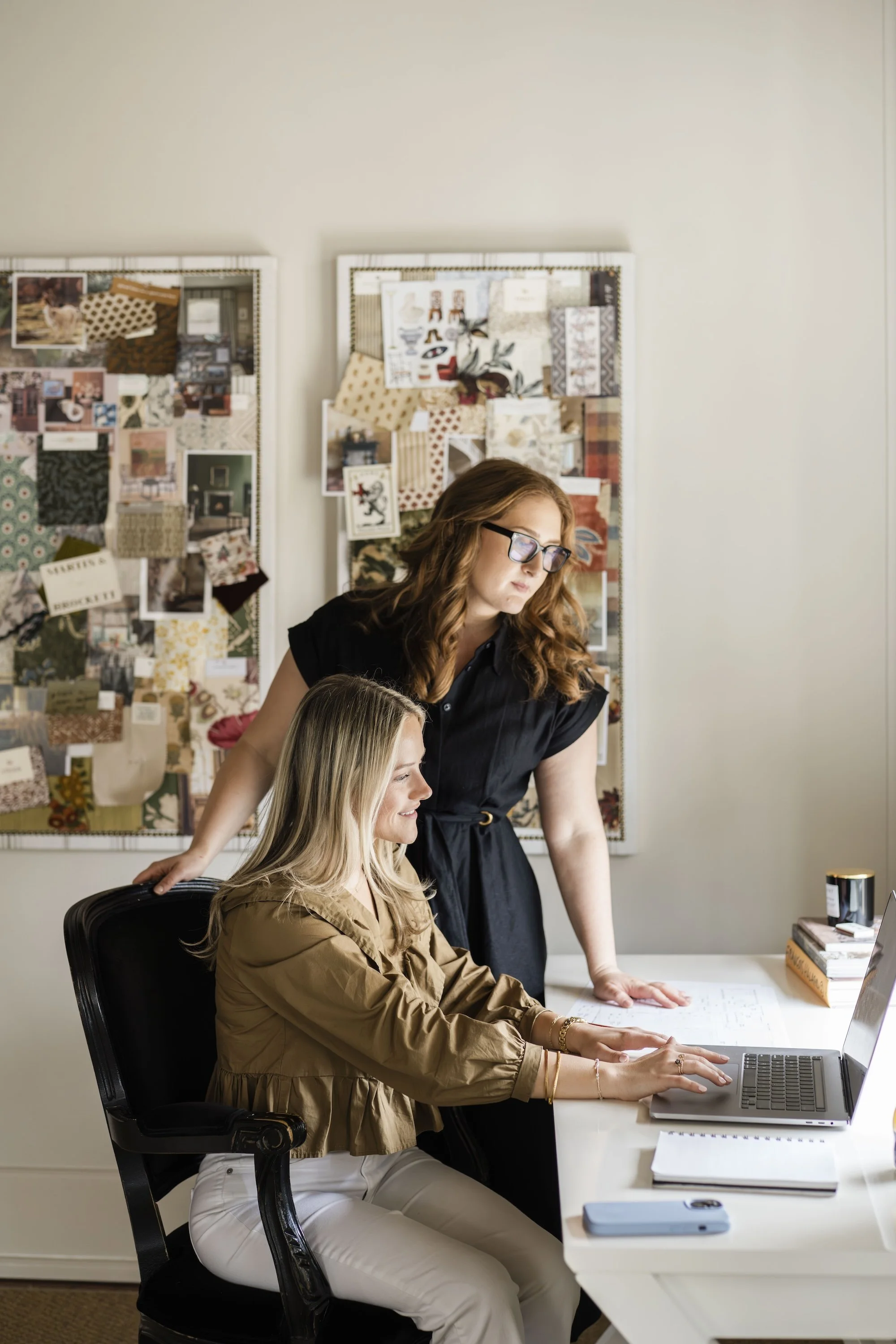 Two women working together at a desk, one seated and the other standing, in an office with bulletin boards filled with fabric swatches and design plans in the background.