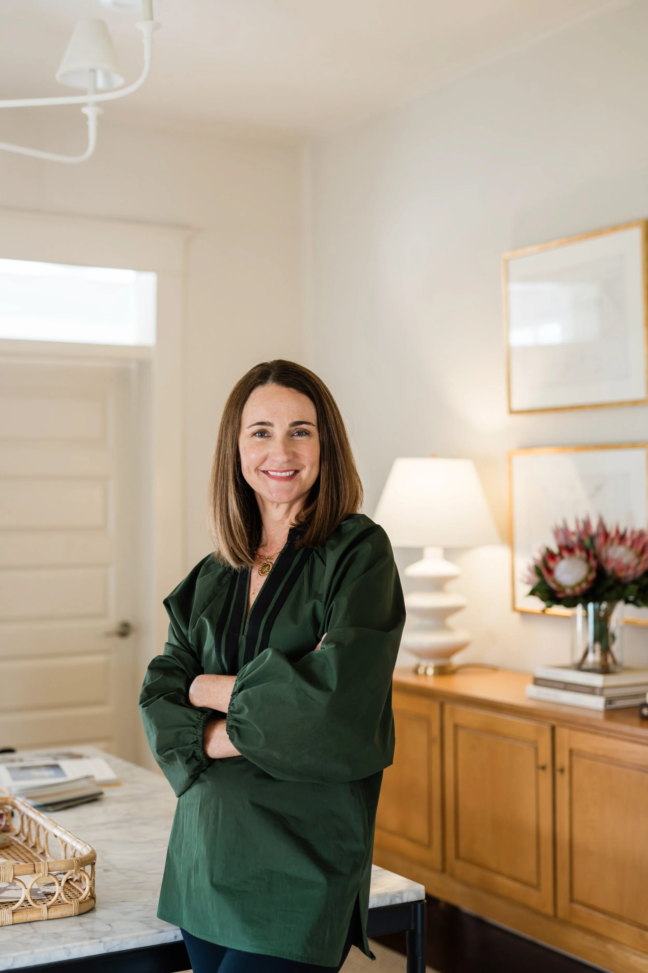 A smiling woman with shoulder-length brown hair, wearing a dark green jacket, standing in a well-lit room with a white wall, wooden sideboard, lamps, and framed pictures.