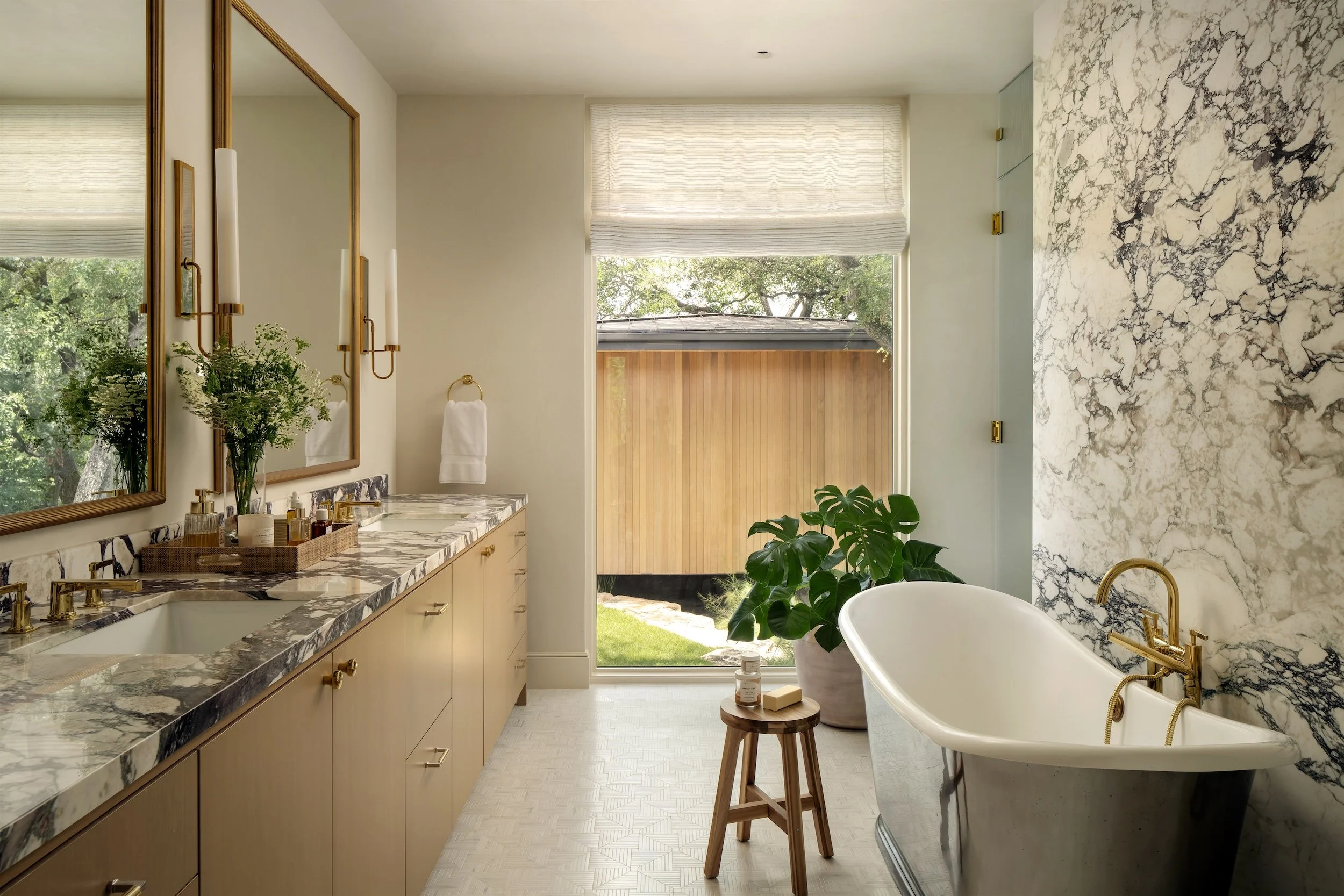 Modern bathroom with a marble countertop double vanity, large mirror, wall-mounted candle sconces, a white towel, a large window with sheer blind, a potted plant, a freestanding bathtub with gold fixtures, and a wooden stool with toiletries.