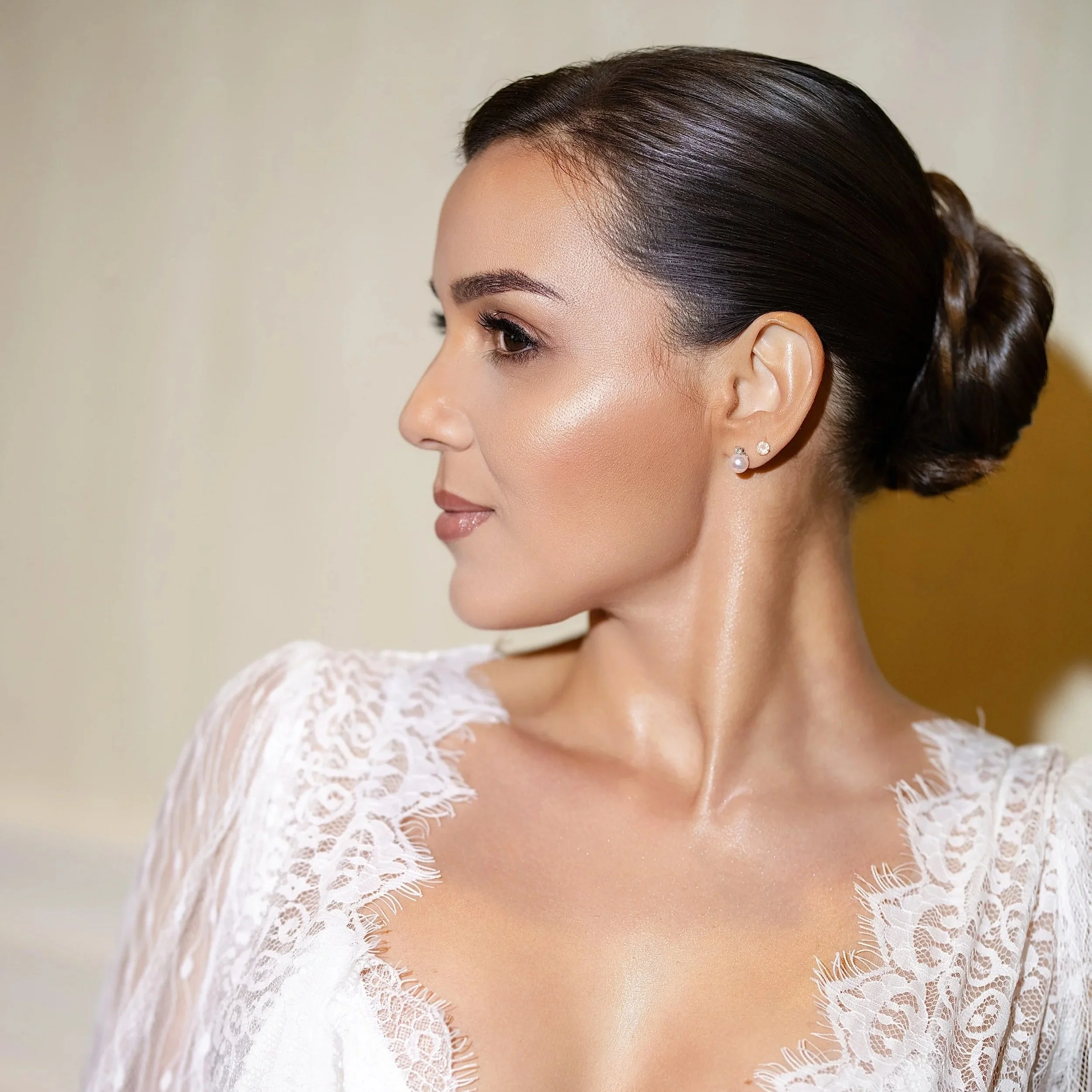 Close-up of a woman's face, showing her dark brown hair styled in an elegant updo, pearl earrings, and wearing a white lace dress.