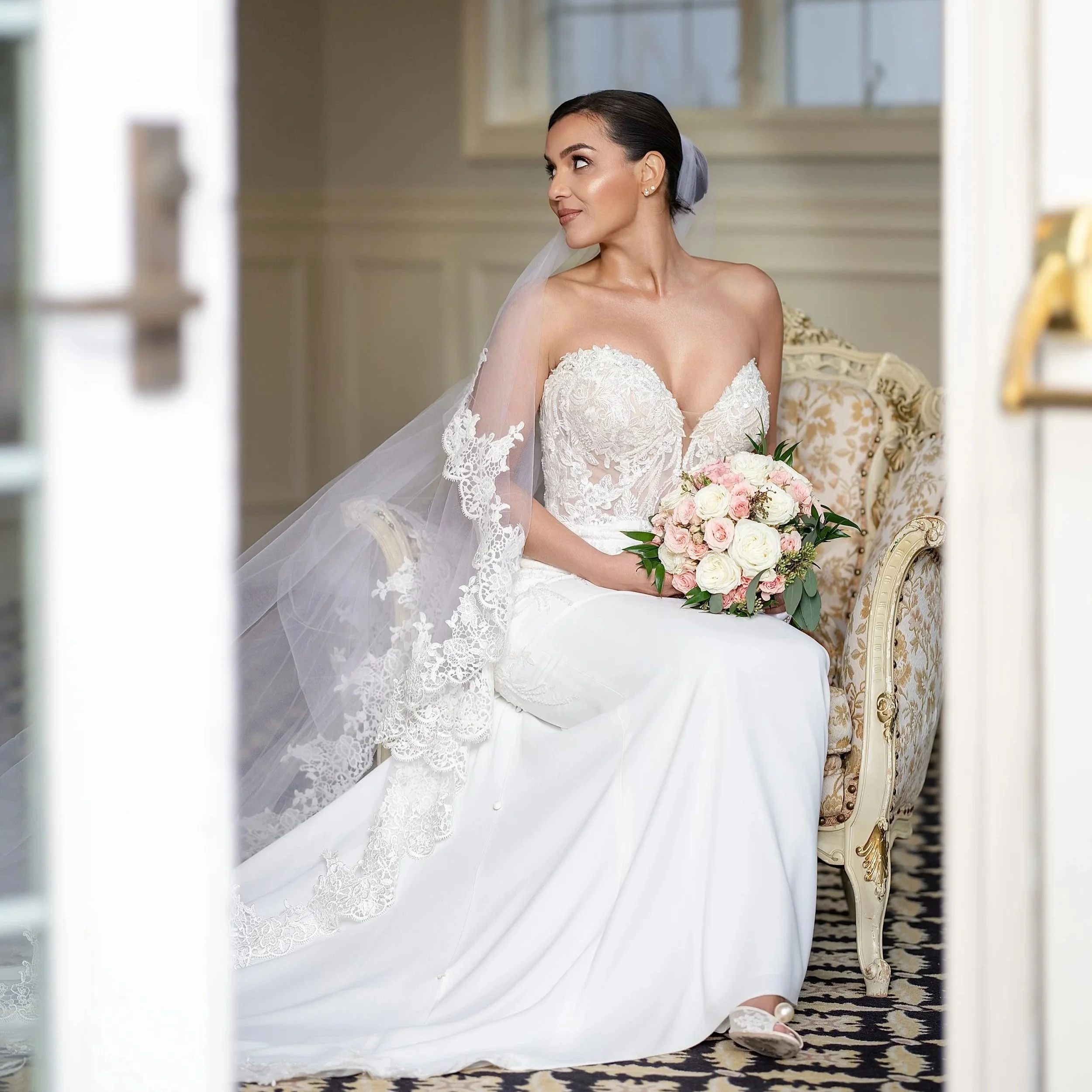 A bride sitting on an ornate chair, holding a bouquet of pink and white roses, wearing a lace wedding dress with a veil, and looking to her right.