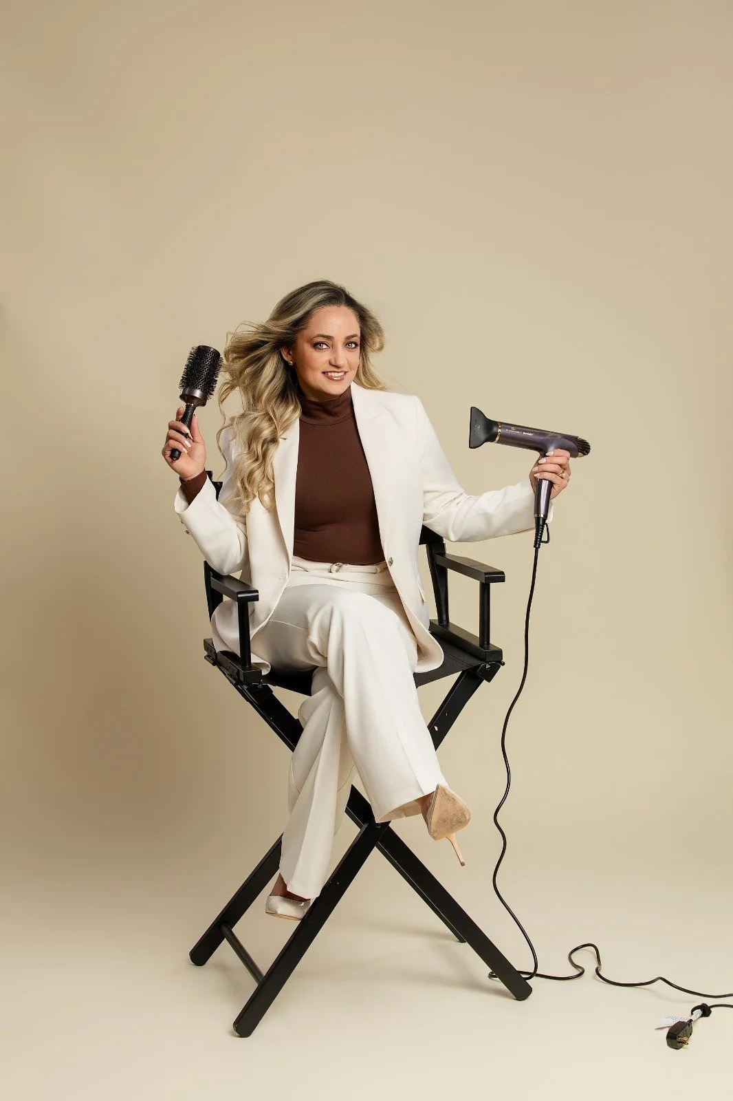 Woman sitting on a director's chair, holding a round hairbrush in one hand and a blow dryer in the other, dressed in a white suit and beige high heels, with a neutral background.