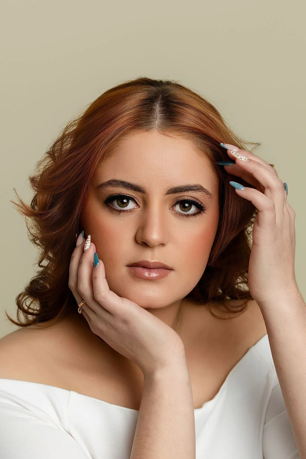 Portrait of a young woman with shoulder-length auburn hair, posed with hands near her face, wearing a white off-shoulder top, minimal makeup, neutral background.