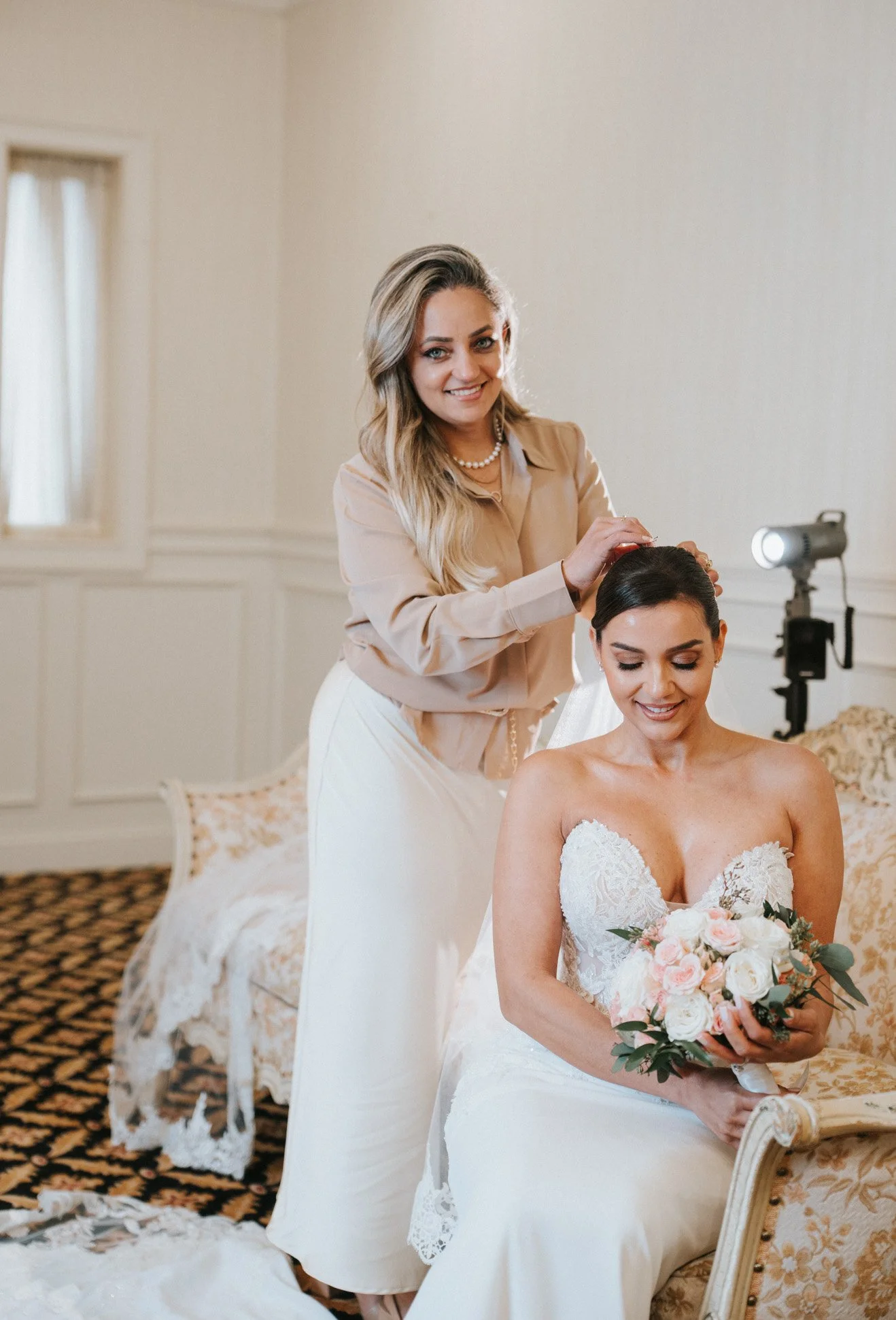 A bride sitting on a chair holding a bouquet of pink and white flowers. A woman standing behind her, smiling, with her hand on the bride's head, in a well-lit room with beige walls, windows, and floral-patterned furniture.