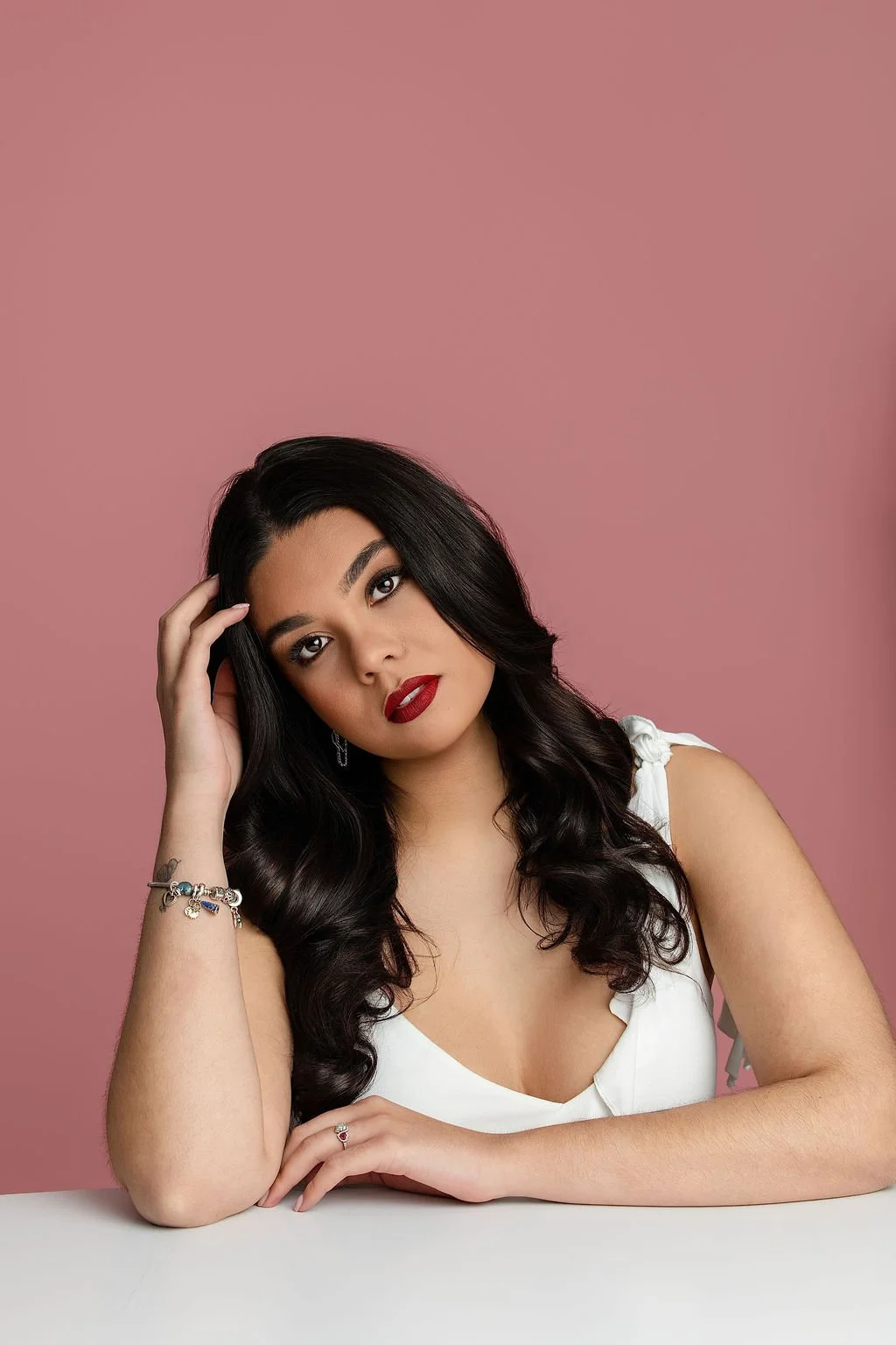 A woman with long dark wavy hair and red lipstick, wearing a white sleeveless top, is sitting at a white table against a pink background. She is resting her head on her left hand, with an expression of contemplation.