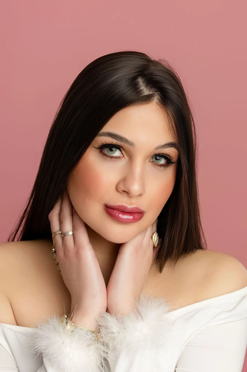 Portrait of a woman with dark brown hair, green eyes, and red lipstick, posing against a pink background, wearing jewelry and an off-shoulder white garment with feather accents.