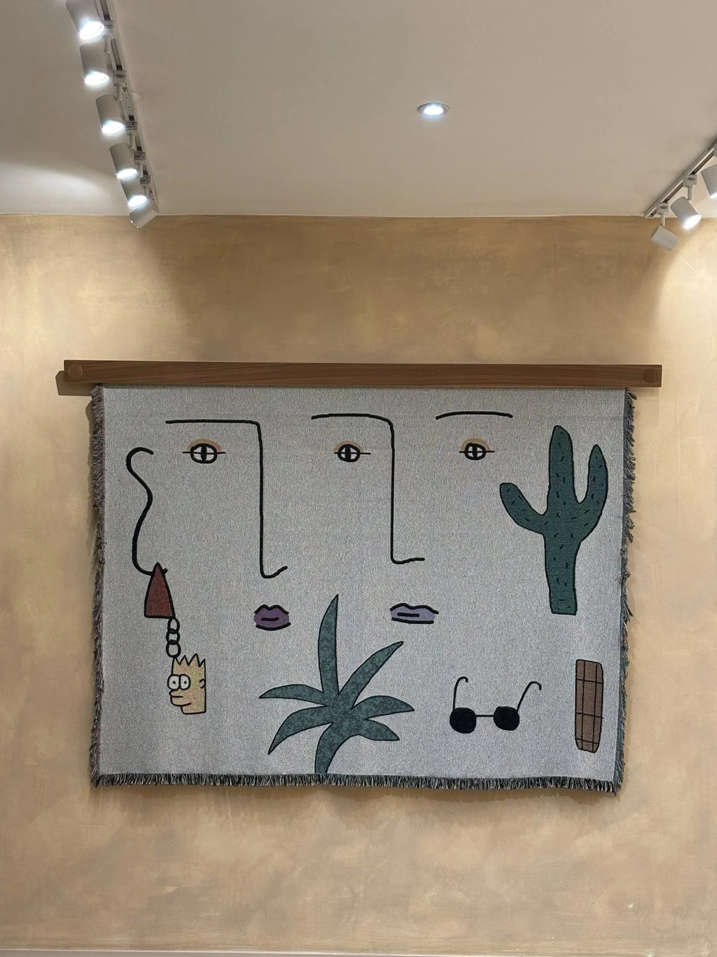 A wall hanging or tapestry with abstract line art faces, cacti, sunglasses, and plant illustrations, mounted on a wooden rod against a beige wall.