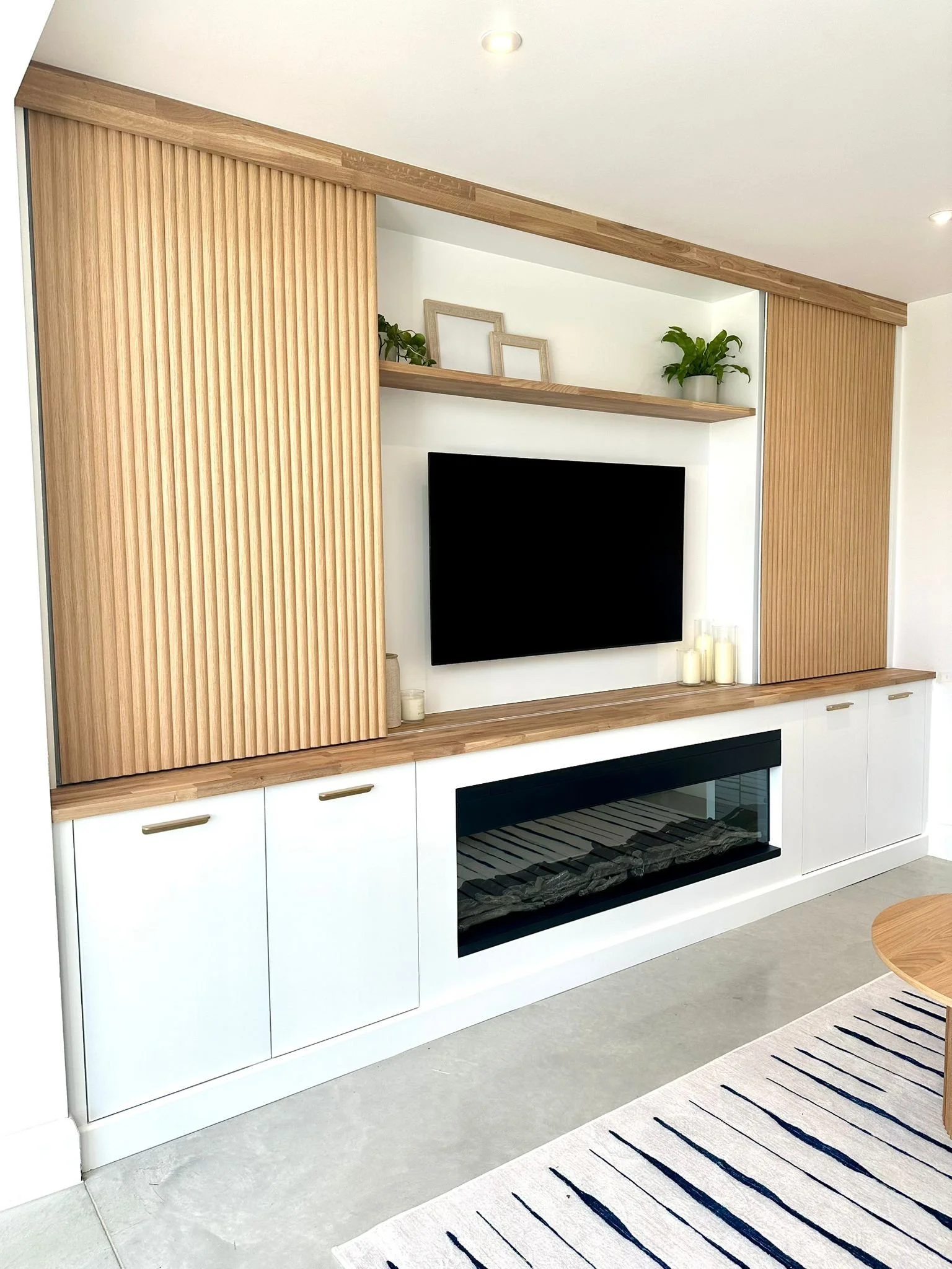 Modern living room entertainment center with a mounted flat-screen TV, a built-in electric fireplace, wooden panel accents on the wall, small decorative candles, and a beige rug with black stripes.