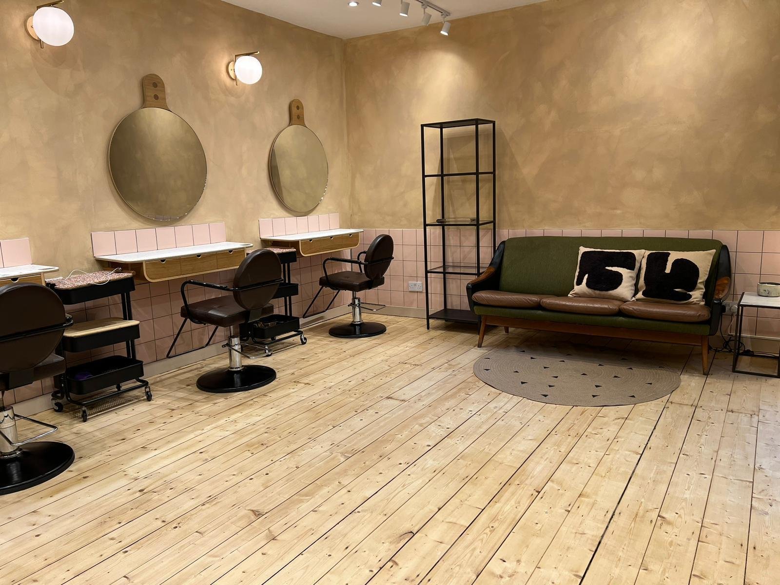 Empty hair salon with three styling stations, large round mirrors, black swivel chairs, a green couch with two black and white pillows, a small oval rug, and a black metal shelving unit against a beige and pink tiled wall.