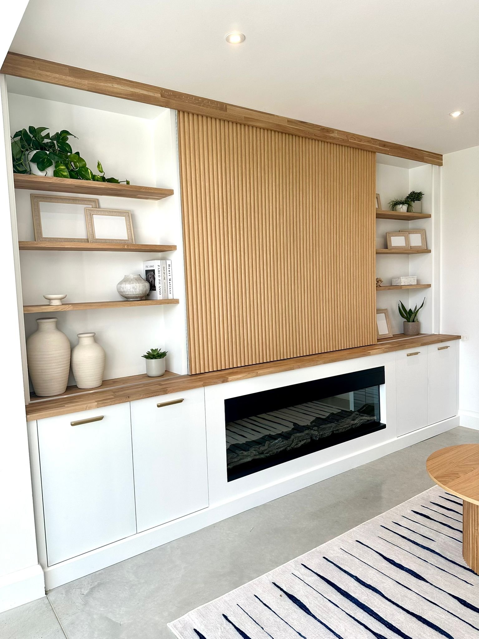 Modern living room with white cabinet, wooden slat accent wall, and open shelves decorated with plants, vases, and picture frames.