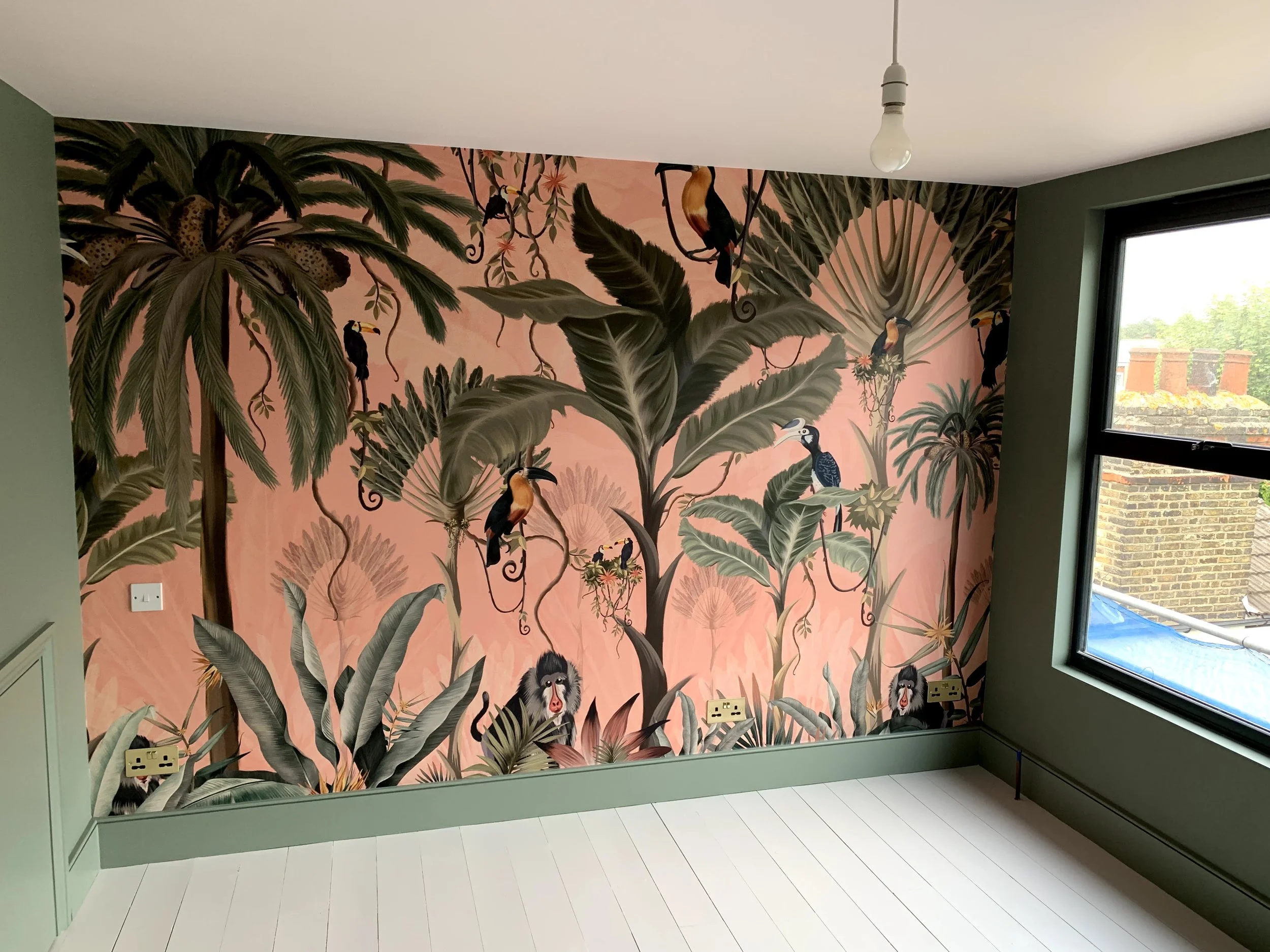 A jungle-themed mural featuring large tropical leaves, monkeys, toucans, and other tropical birds on a pink background, with a window to the right showing a brick building and trees outside.