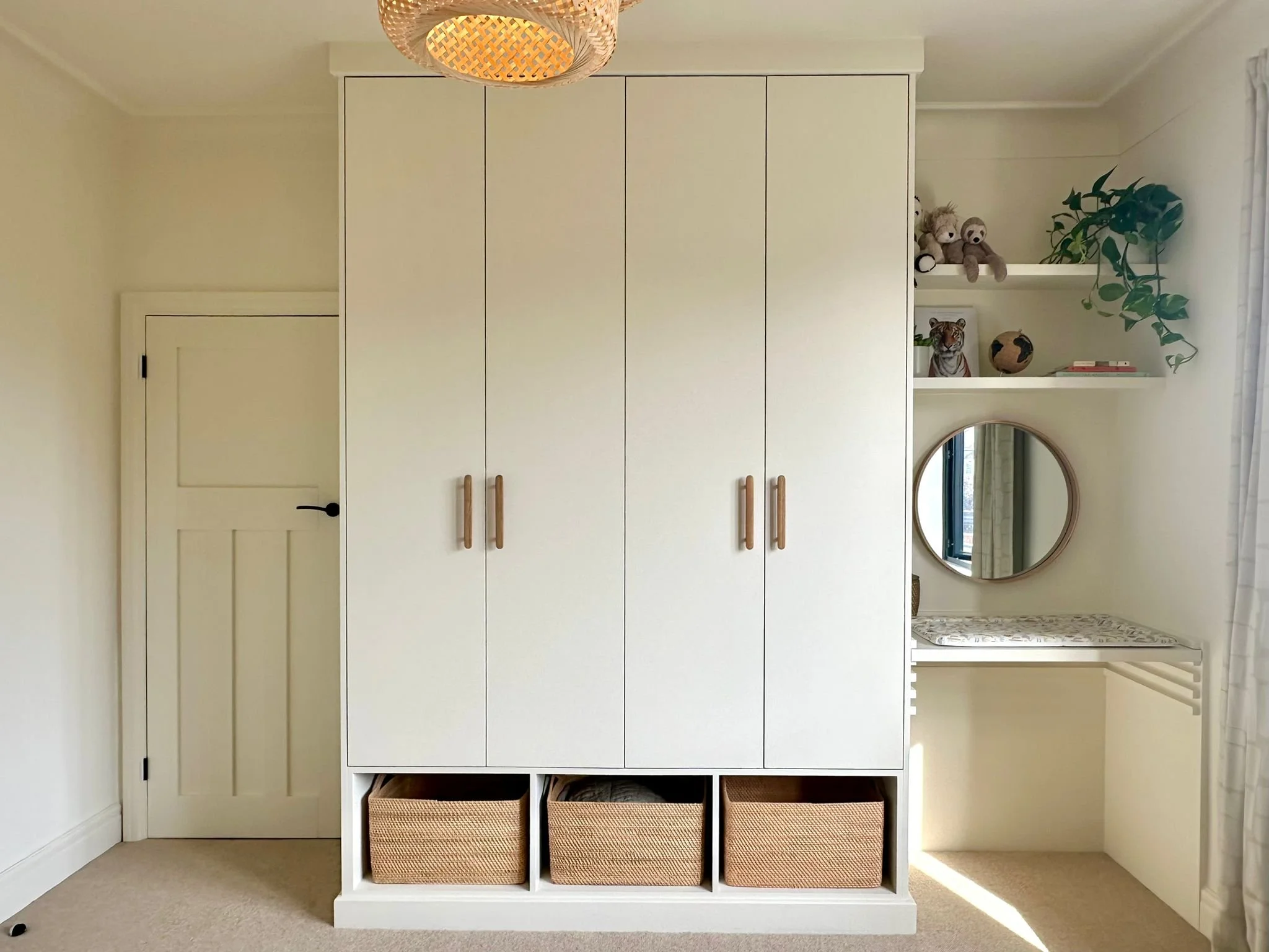 Closet with white doors, wooden handles, and open storage baskets at the bottom, next to a small vanity area with a round mirror, shelves with stuffed animals, a small globe, and a plant, in a light-colored room with beige carpet.