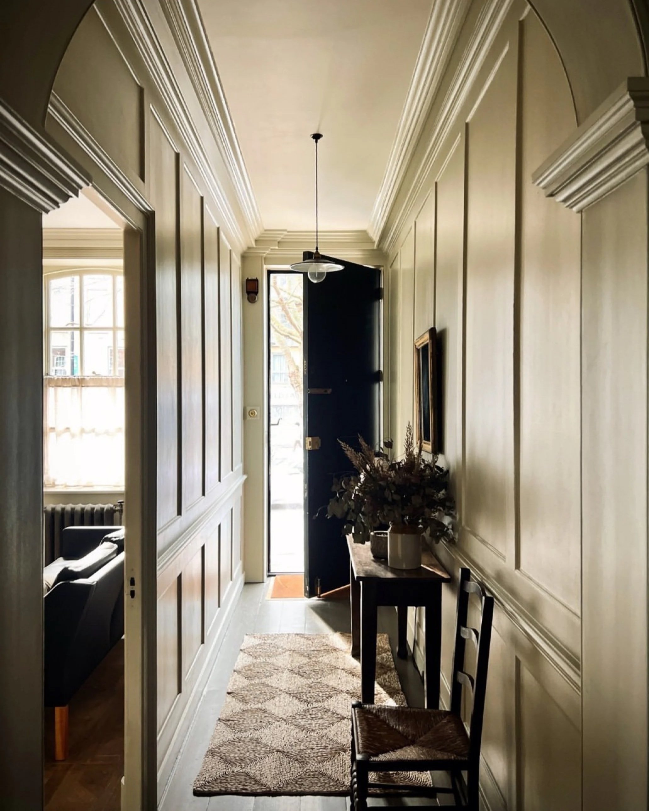 A cozy hallway with wooden wall panels, leading to an open front door allowing sunlight to brighten the space. There's a small table with a floral arrangement, a framed picture on the wall, and two chairs along the right side. A rug runs down the floor, and a glimpse of a living room with a black sofa is visible on the left.