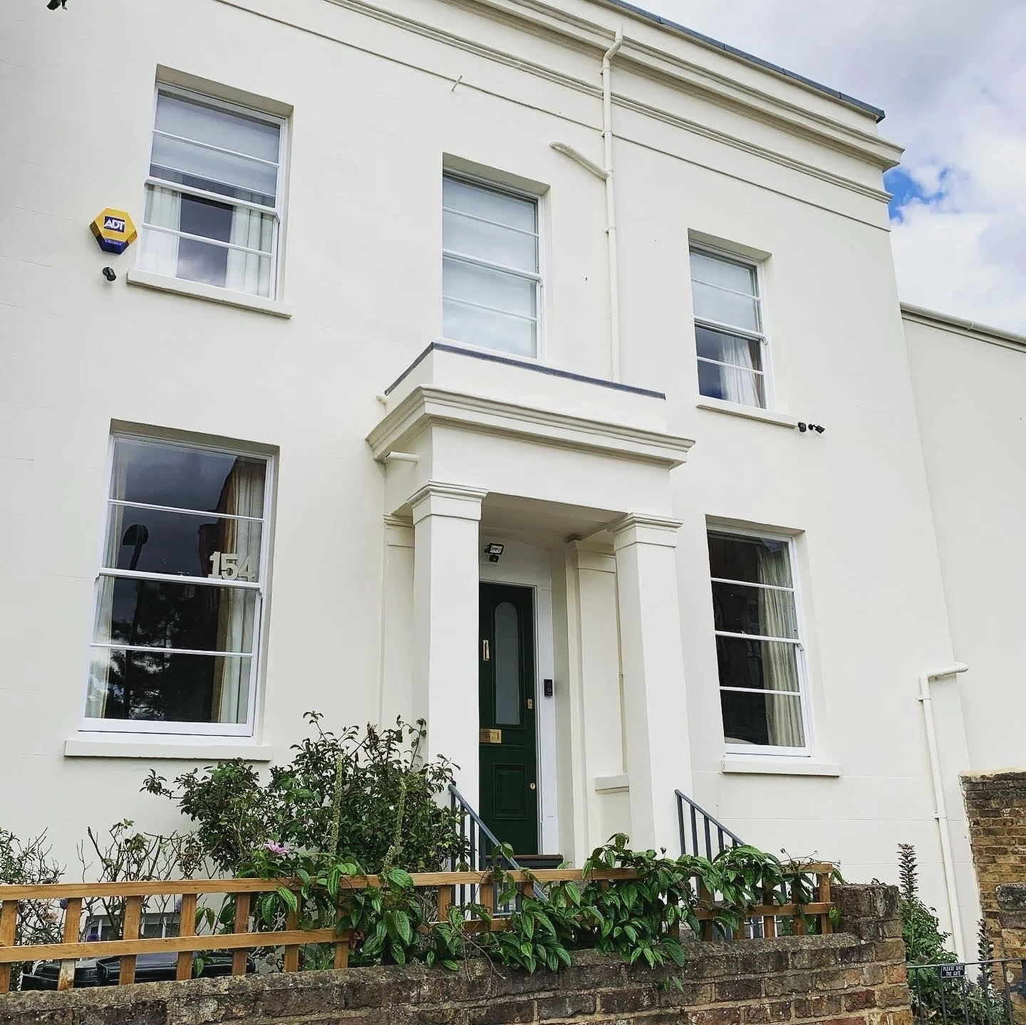 White multi-story house with green front door, large windows, and a small garden with bushes and a brick fence.