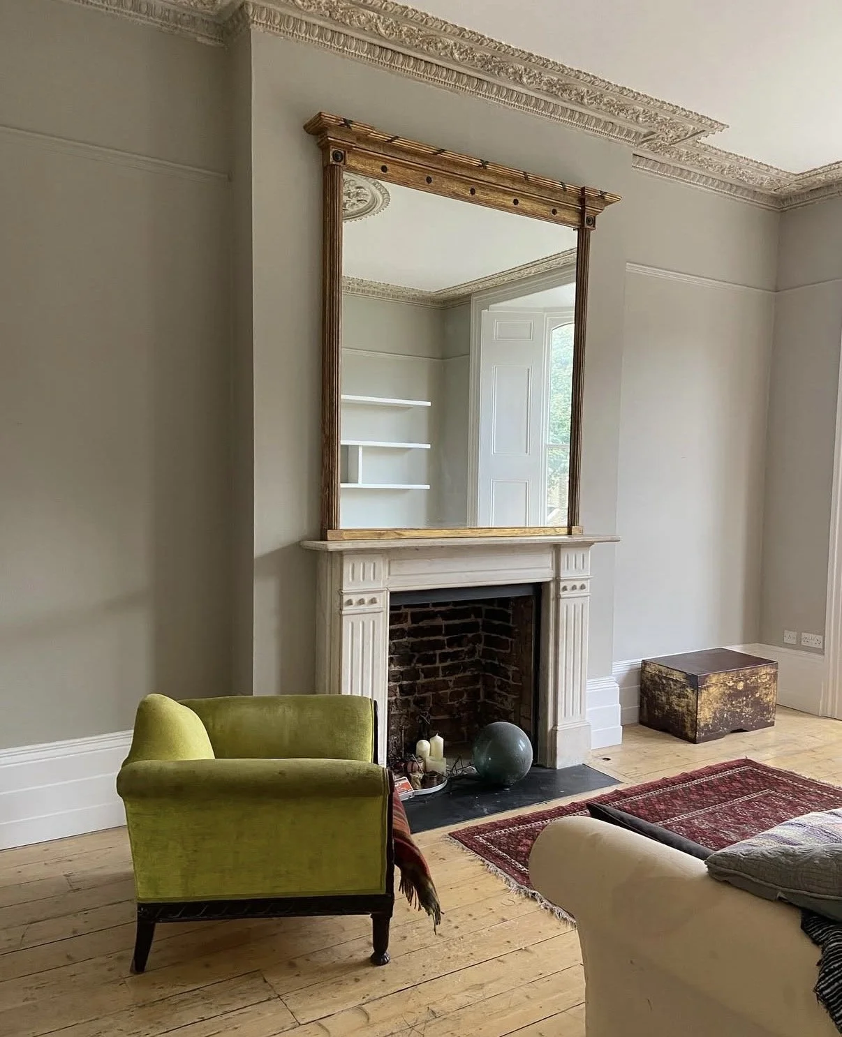 Living room with a fireplace, a large mirror above it, a green armchair, a beige sofa, a red area rug, and a window with white shelves.