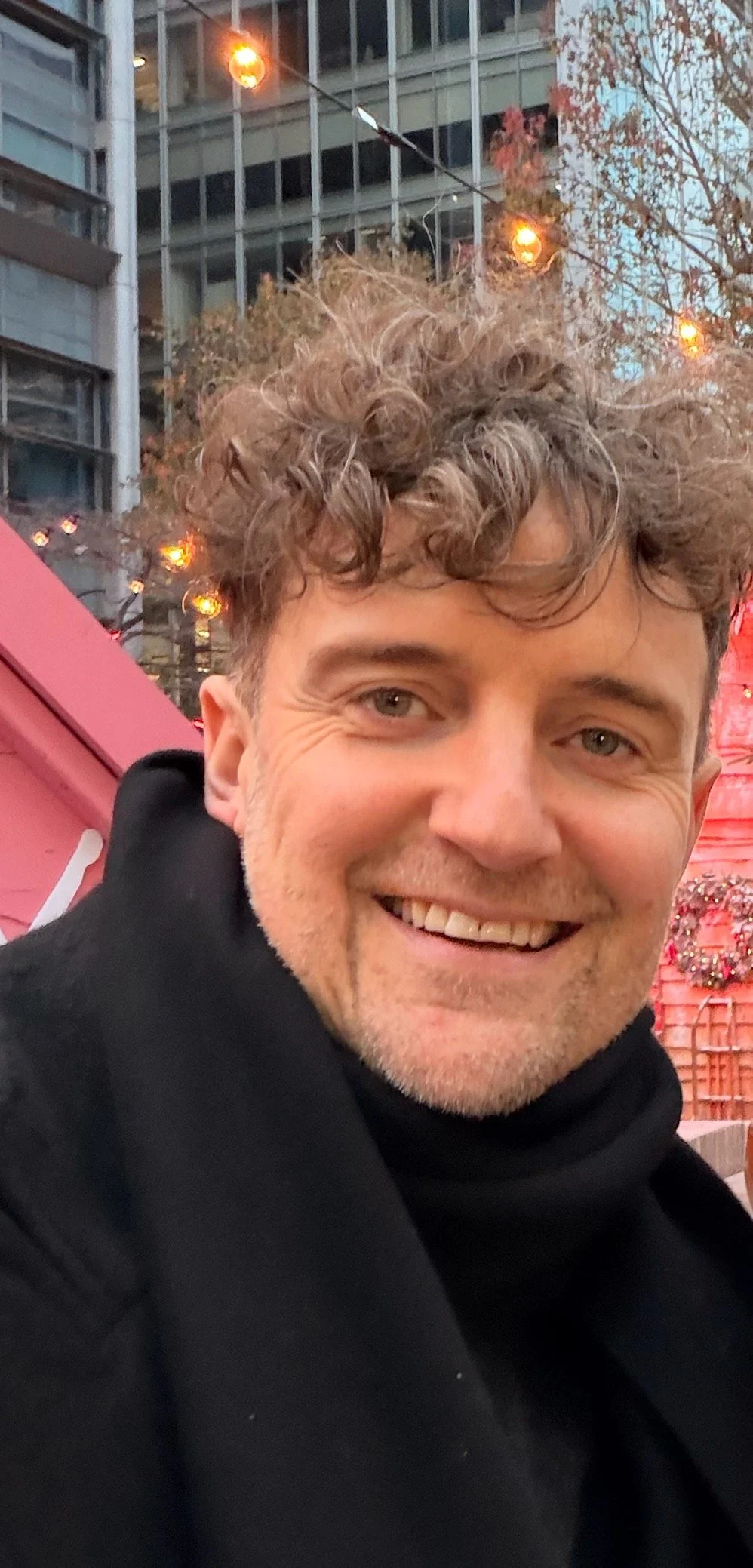 Close-up of a smiling man with curly hair, wearing a black coat outdoors in an urban setting during evening with string lights and a decorated building in the background.
