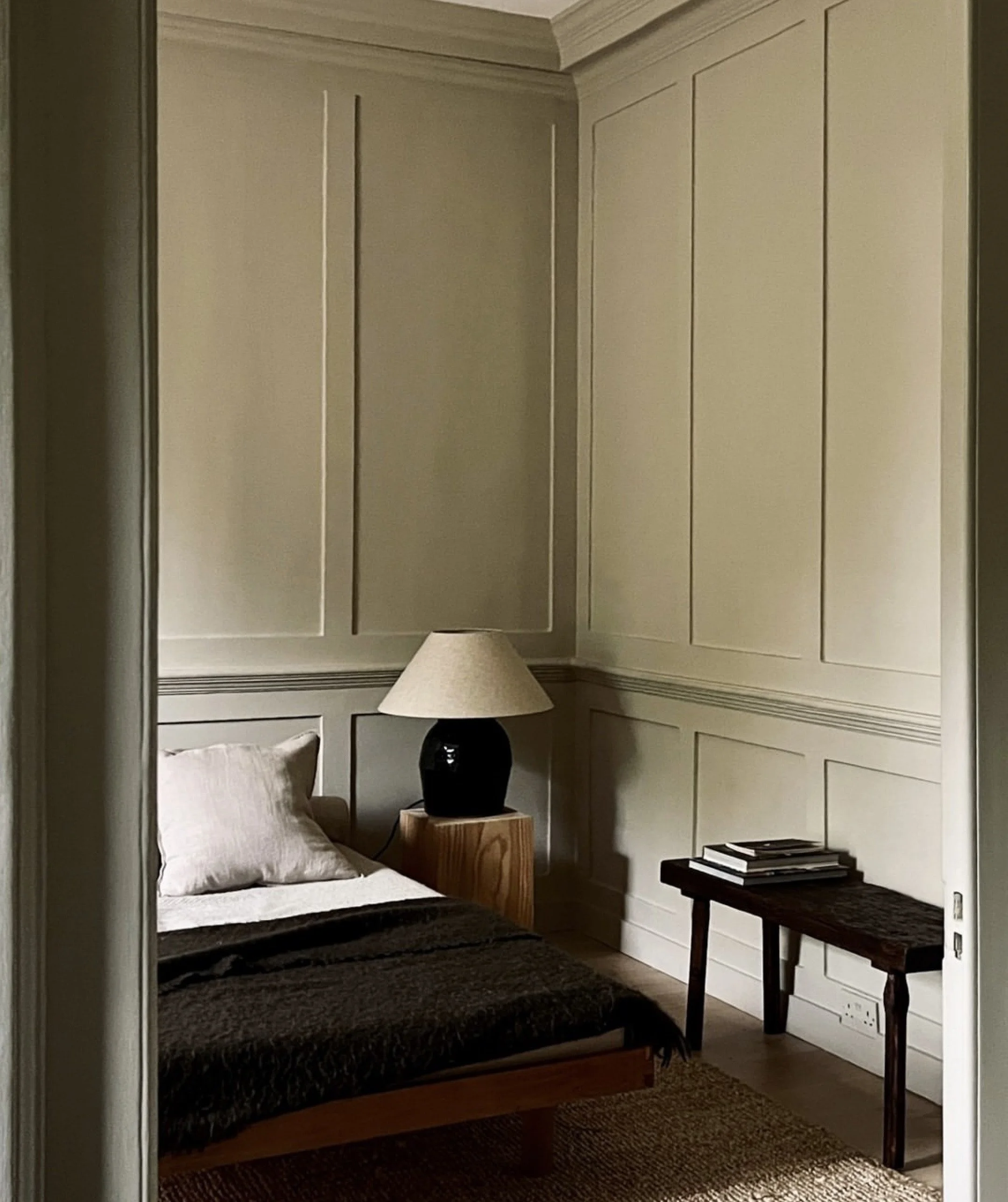 A bedroom with cream-colored panel walls, a wooden bed frame with white pillow and dark blanket, a bedside wooden table with a black lamp and beige lampshade, and a small dark wooden table with some books on it, next to a power outlet and woven rug on the floor.