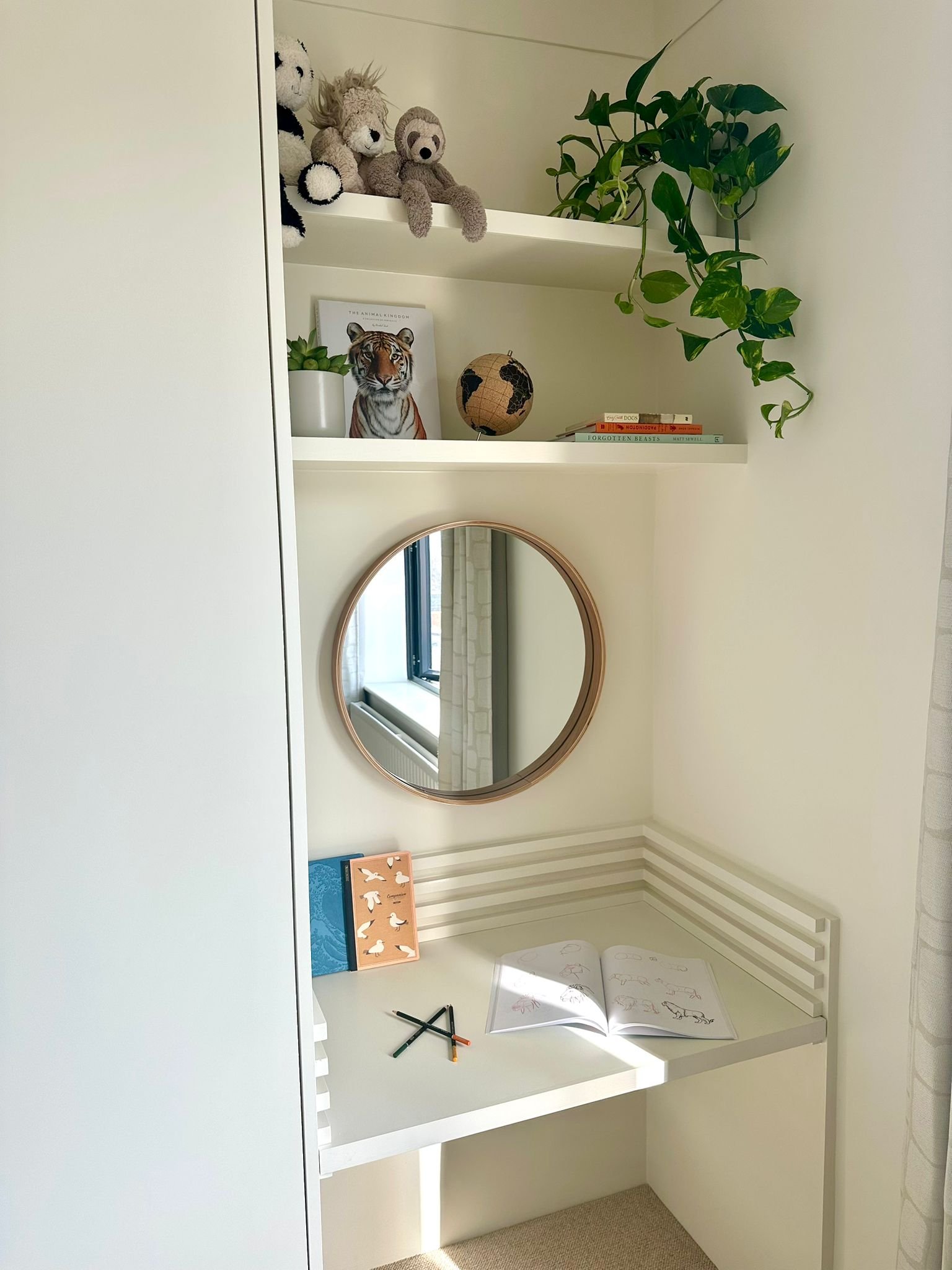 A white corner desk with an open sketchbook, some pens, and books on it. Above it, a round mirror on the wall reflects a window with curtains. There are two white shelves above the desk, holding plush toys, a potted plant, a framed animal picture, a small globe, and some books. A hanging green plant cascades from the top shelf.