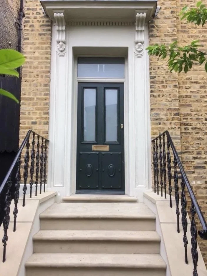 Black front door with trim and a mail slot, accessed by three beige stone steps with black metal railings on both sides, beside a brick wall with some green foliage.