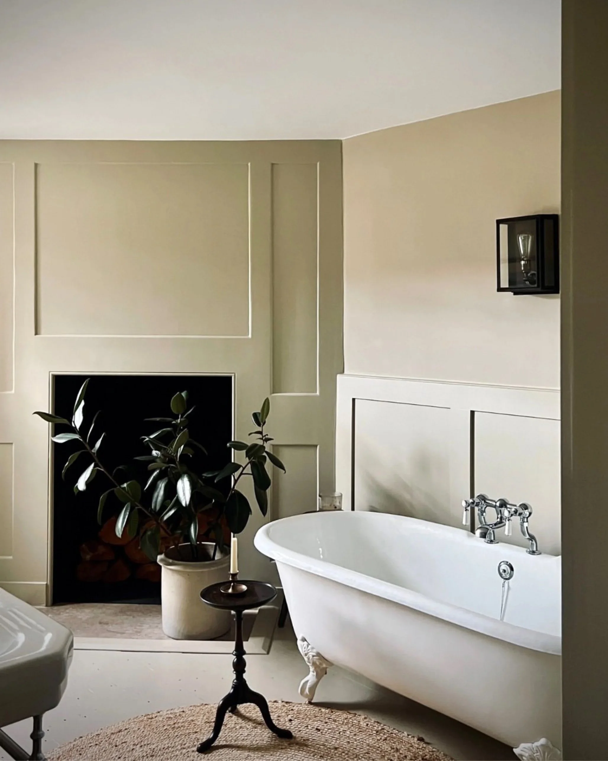A vintage white clawfoot bathtub next to a wall with beige paneling, a black wall-mounted shelf with a decorative light, a large potted plant, a small dark side table with a candle, and a brown circular rug on the floor.