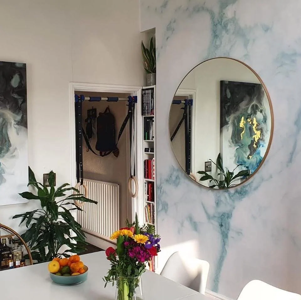 A dining room with a white table, a glass vase of colorful flowers, a bowl of assorted fruits, a large round mirror on a marble-patterned wall, and a bookshelf in the background.