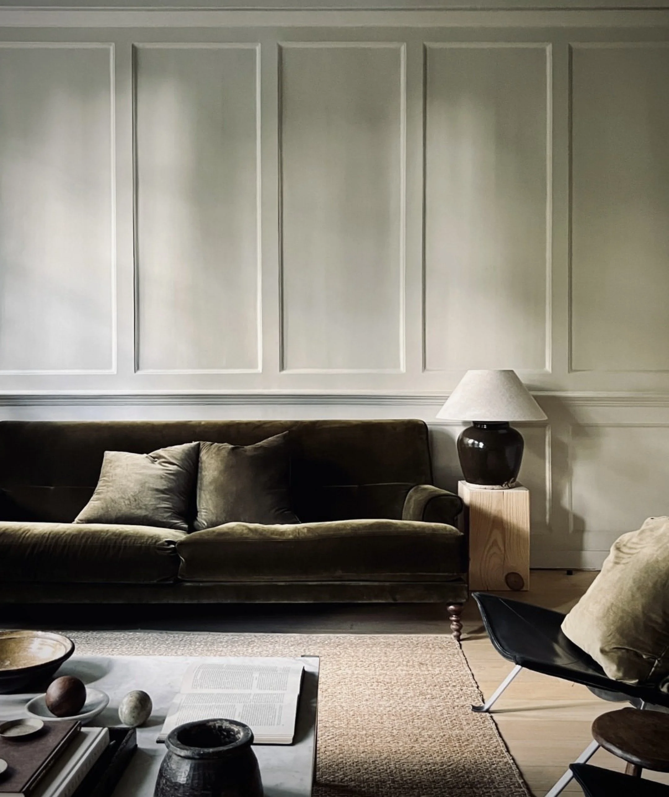 Living room with a brown velvet sofa, beige pillows, a side table with a ceramic lamp, a black lounge chair, a coffee table with books and decorative objects, and a textured area rug.