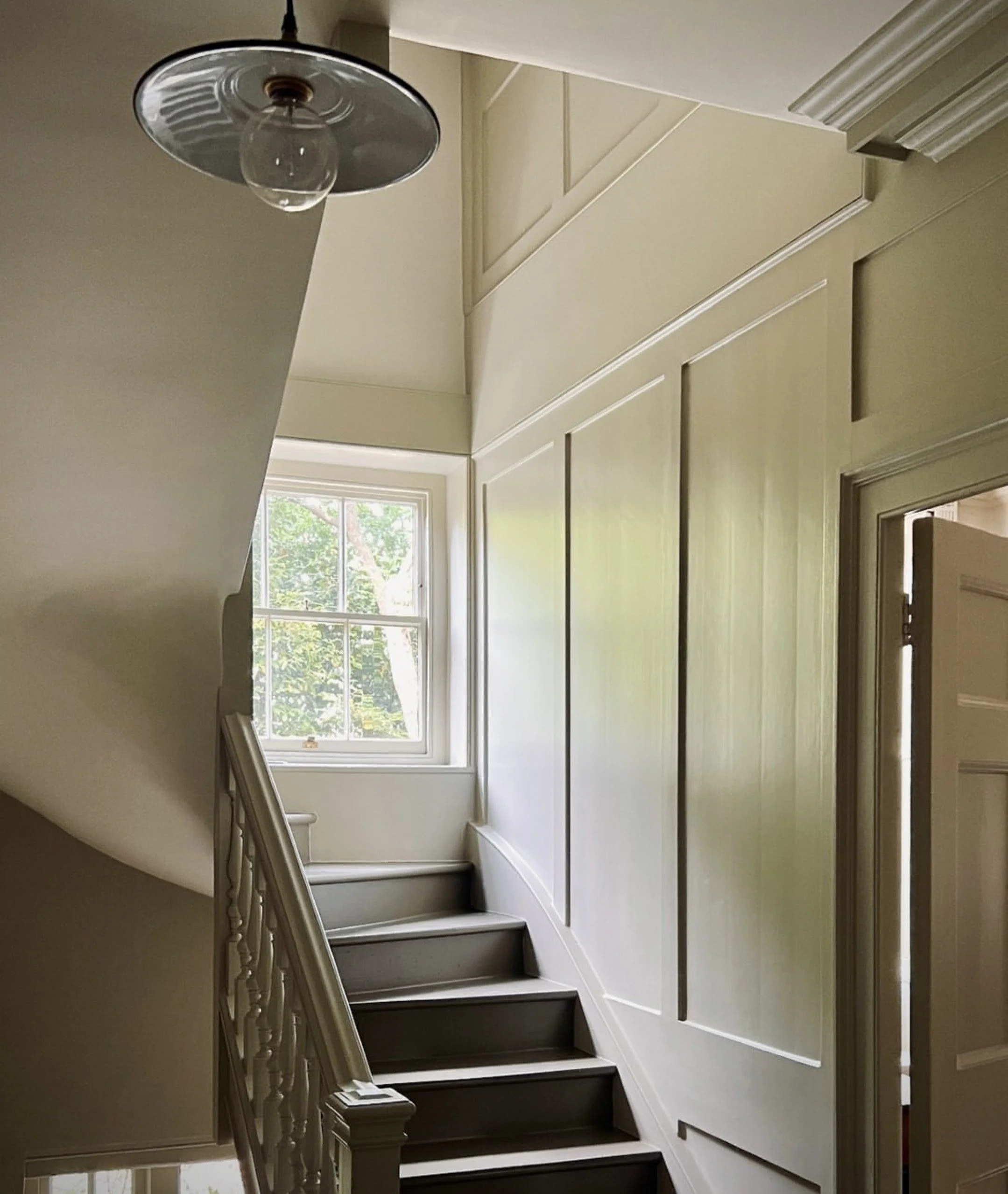 Interior staircase with white walls, window at the top letting in natural light, main staircase with dark steps, decorative paneling on wall, ceiling light fixture.