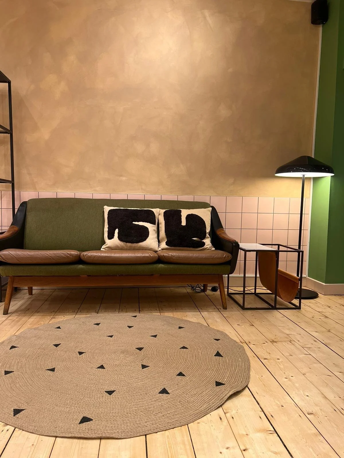 A living room with a green sofa, two pillows with black numbers, a beige circular rug with black triangles, a wooden floor, a black floor lamp with a round shade, and a side table with a glass top and metal frame.