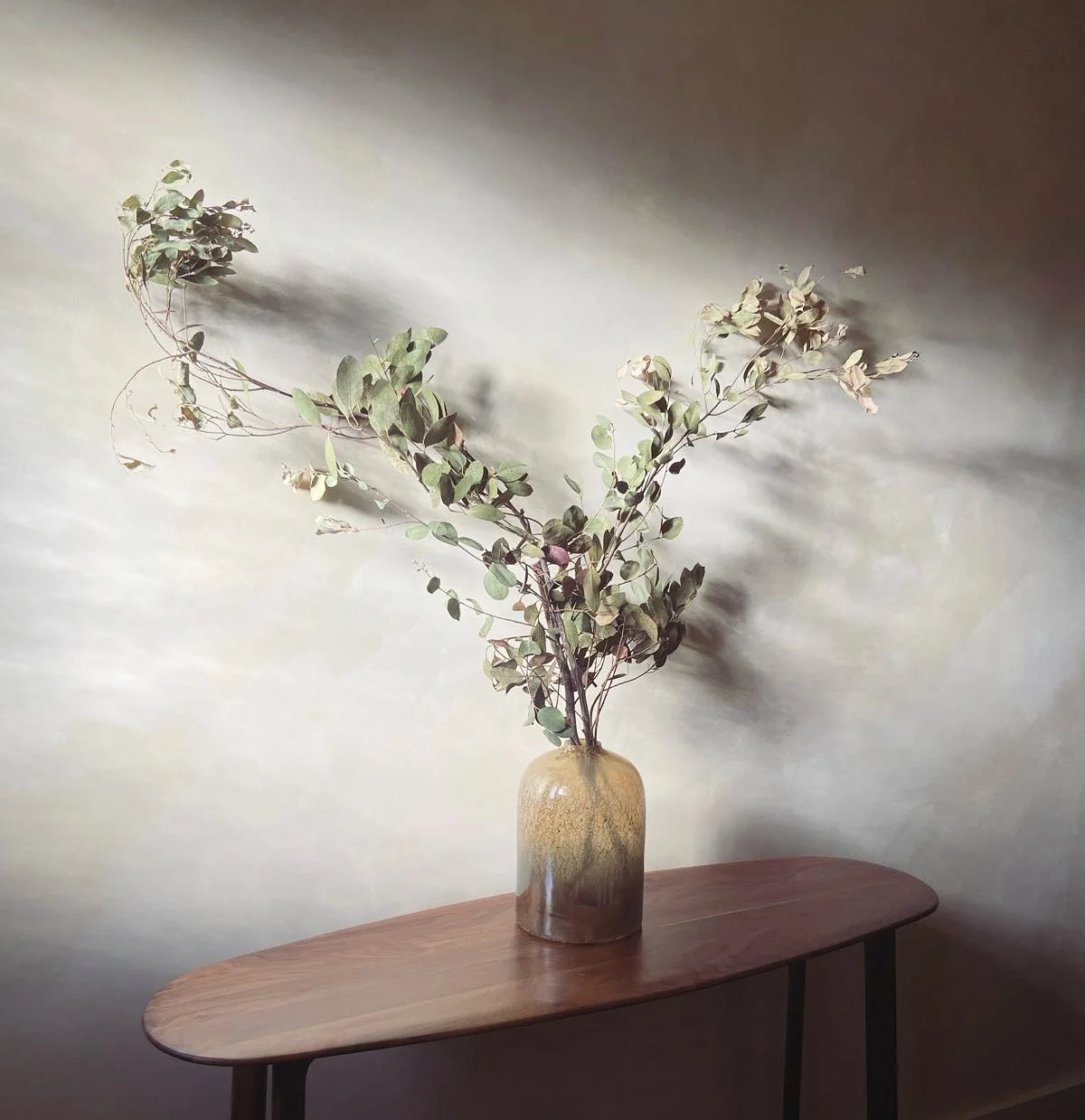 A tall, brown ceramic vase with dried eucalyptus branches sits on a narrow, wooden table against a neutral wall.