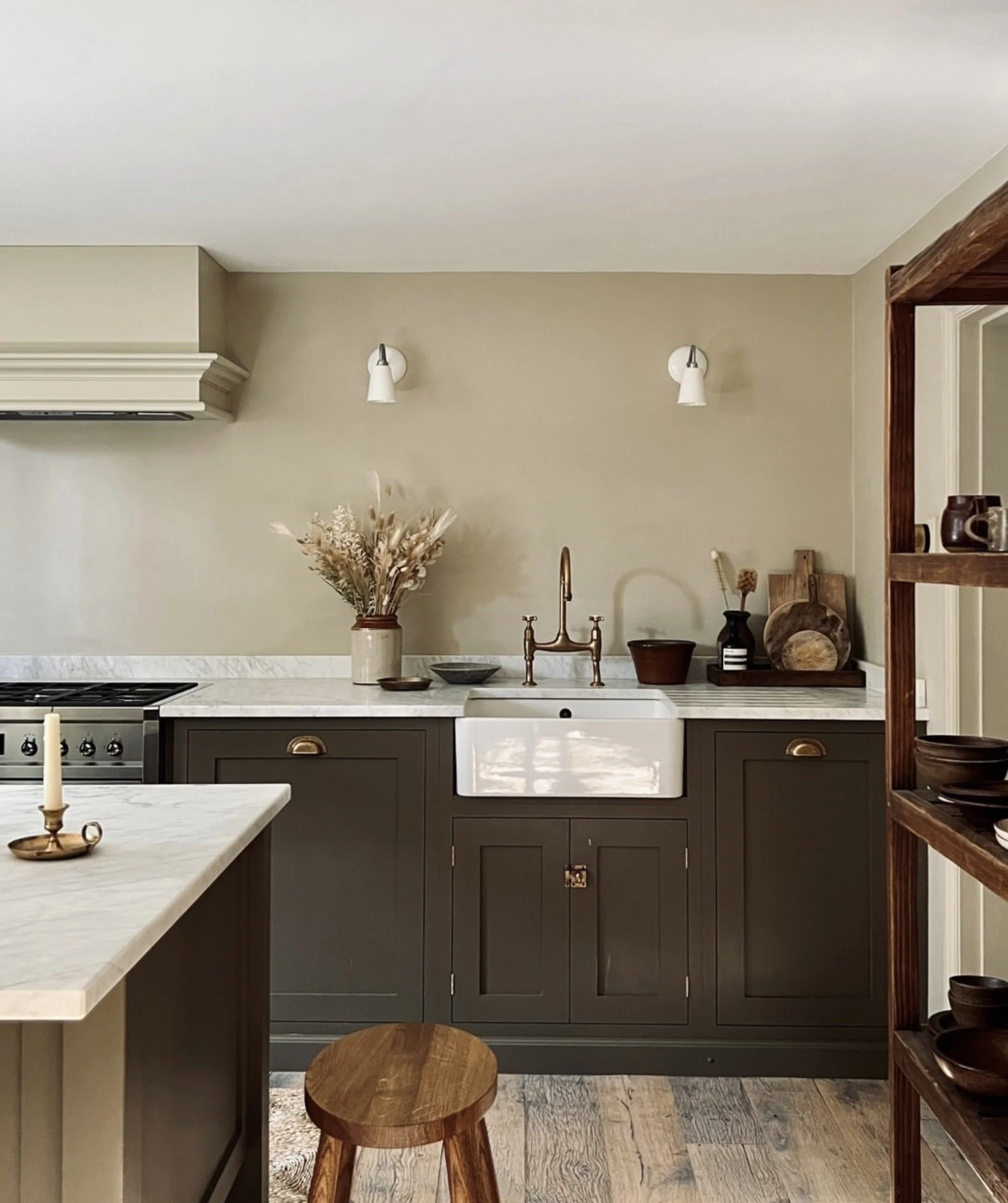 Kitchen with beige walls, white marble countertops, dark green cabinets, and open shelving with dark bowls. A white farmhouse sink with a brass faucet, decorative flowers in a beige vase, and wooden accessories are arranged on the counter. Two white wall sconces above the sink provide lighting, and a wooden stool is in front of the counter.