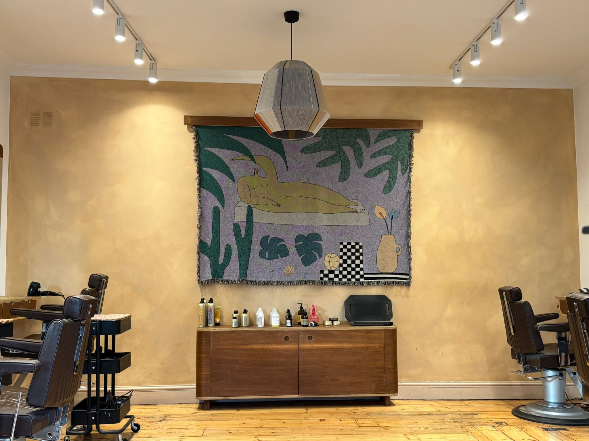 Interior of a barbershop with barber chairs, a wooden cabinet, and a colorful wall hanging of a reclining woman, surrounded by green abstract shapes and vases, under a hanging pendant light.